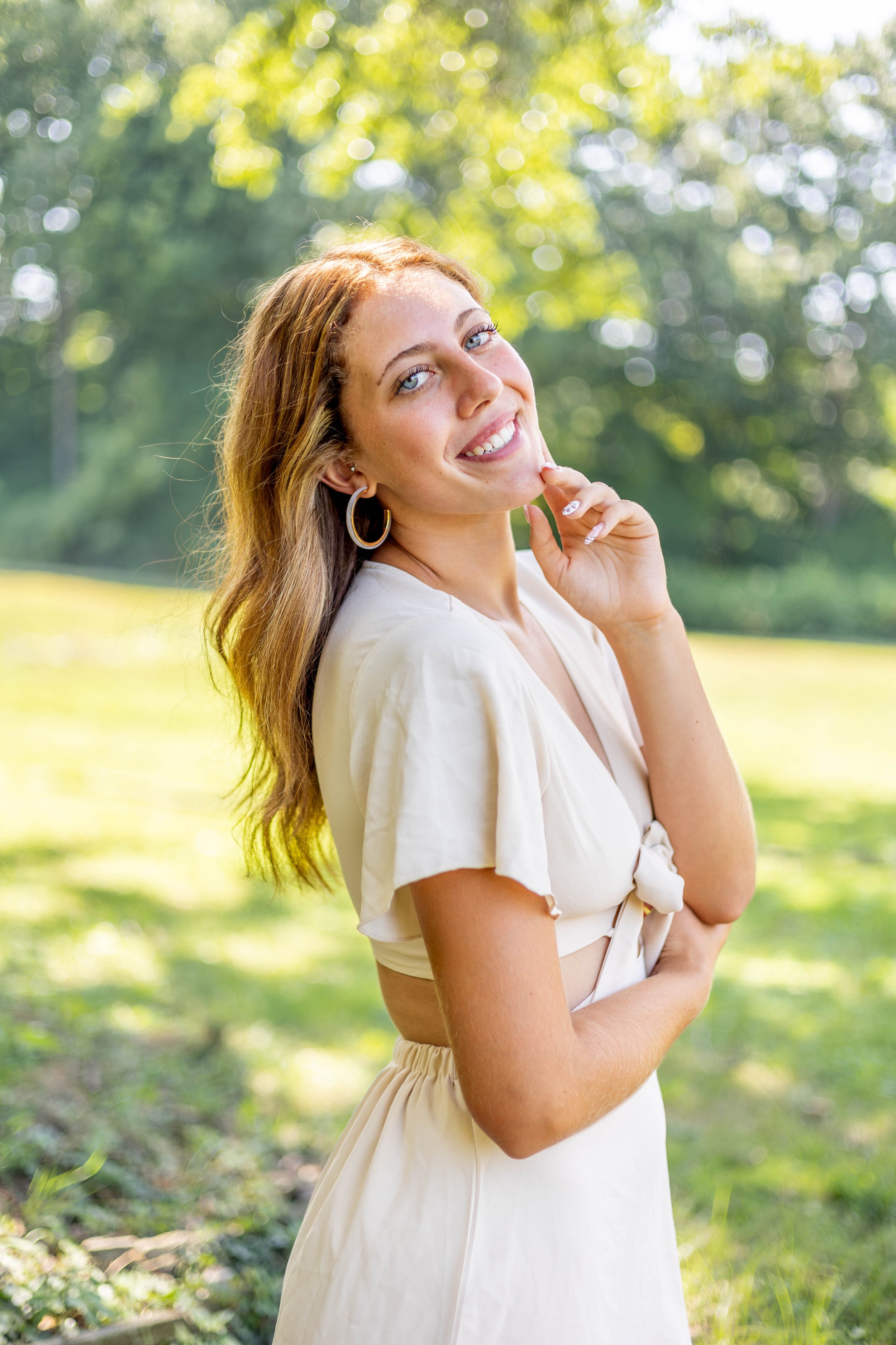 A woman with long reddish hair and blue eyes smiling outdoors on a sunny day with a blurred background of trees and grass.