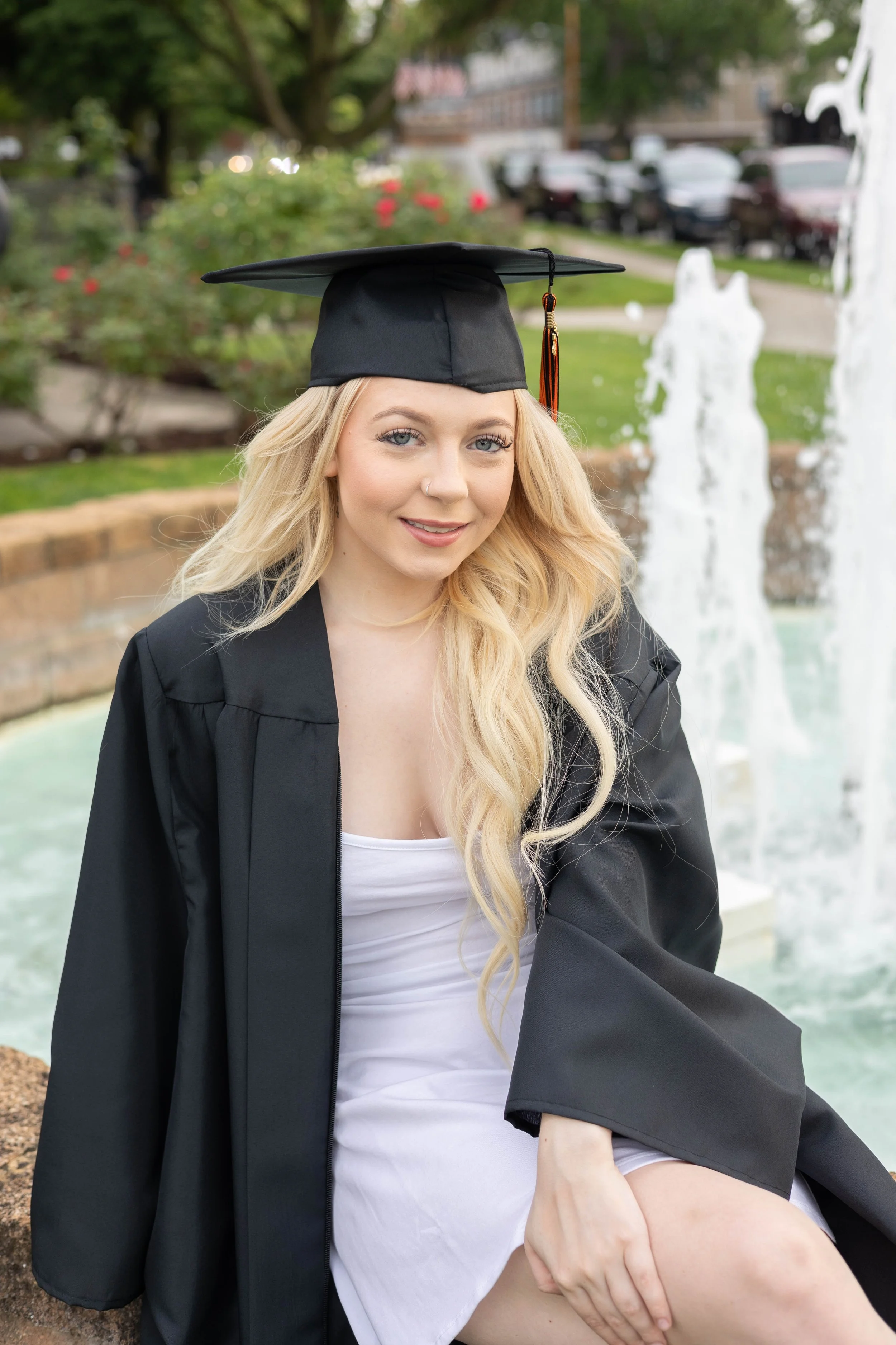 A young woman in a graduation gown and cap sitting outdoors near a fountain with water streams, with trees and parked cars in the background.