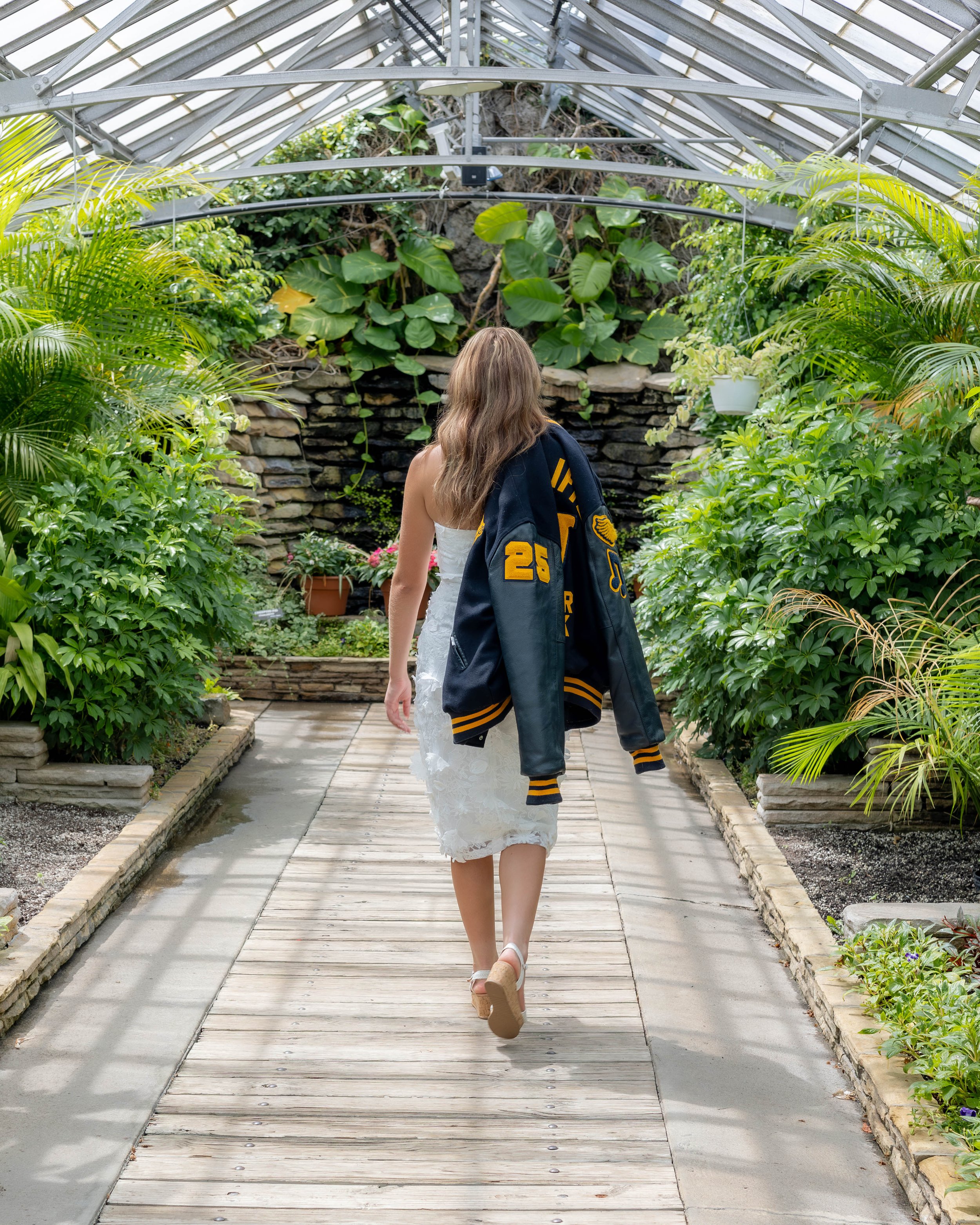 A woman walking away in a greenhouse with lush green plants on either side of a wooden pathway.