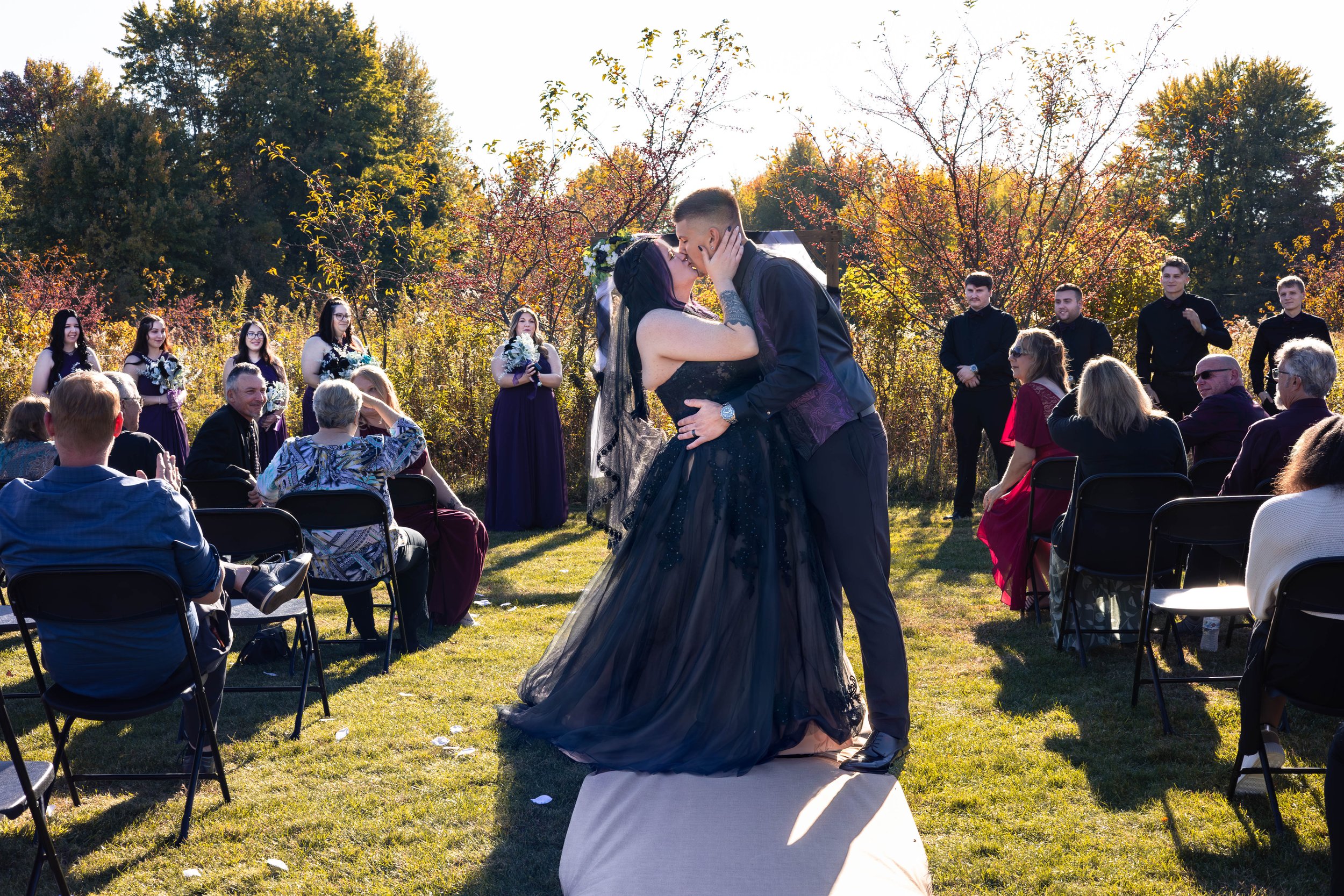 Couple shares a kiss during a wedding ceremony outdoors with guests watching and colorful trees in the background on a sunny day.