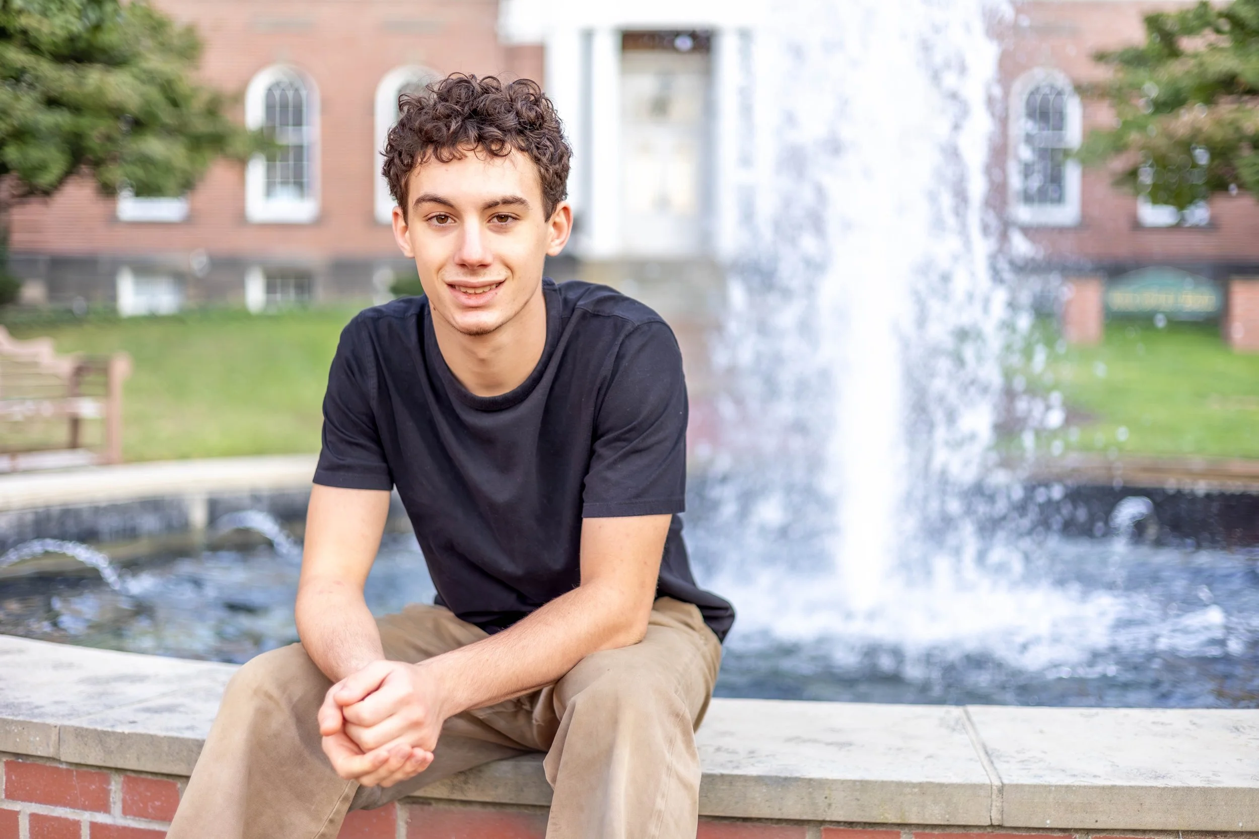 A young man with curly hair sitting outdoors in front of a fountain and a brick building with arched windows.