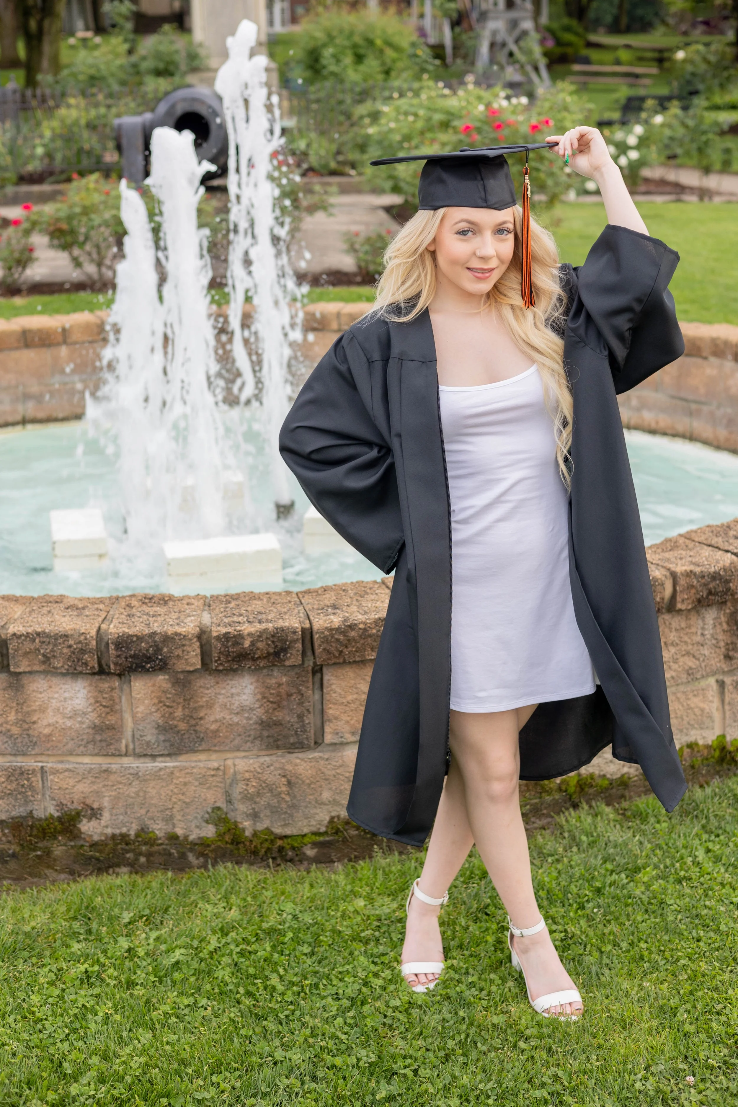 A young woman in a black graduation gown and cap, with a white dress underneath, standing outdoors near a fountain, adjusting her cap with her right hand. She has long blonde hair and is wearing white high-heeled sandals, smiling at the camera with a