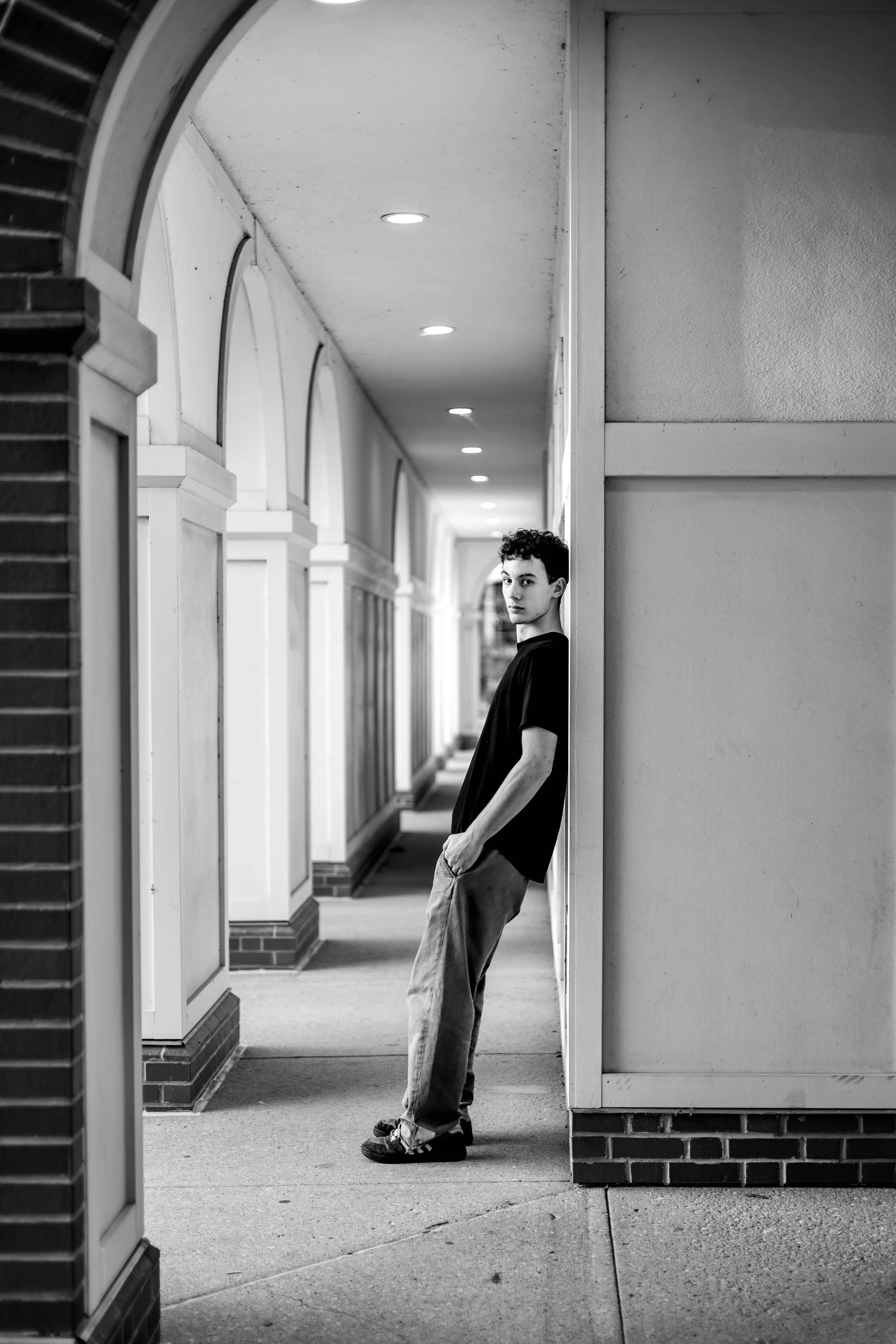A young man with dark curly hair, wearing a black T-shirt and light jeans, leaning against a wall outside a building with arches, on a sidewalk at night.