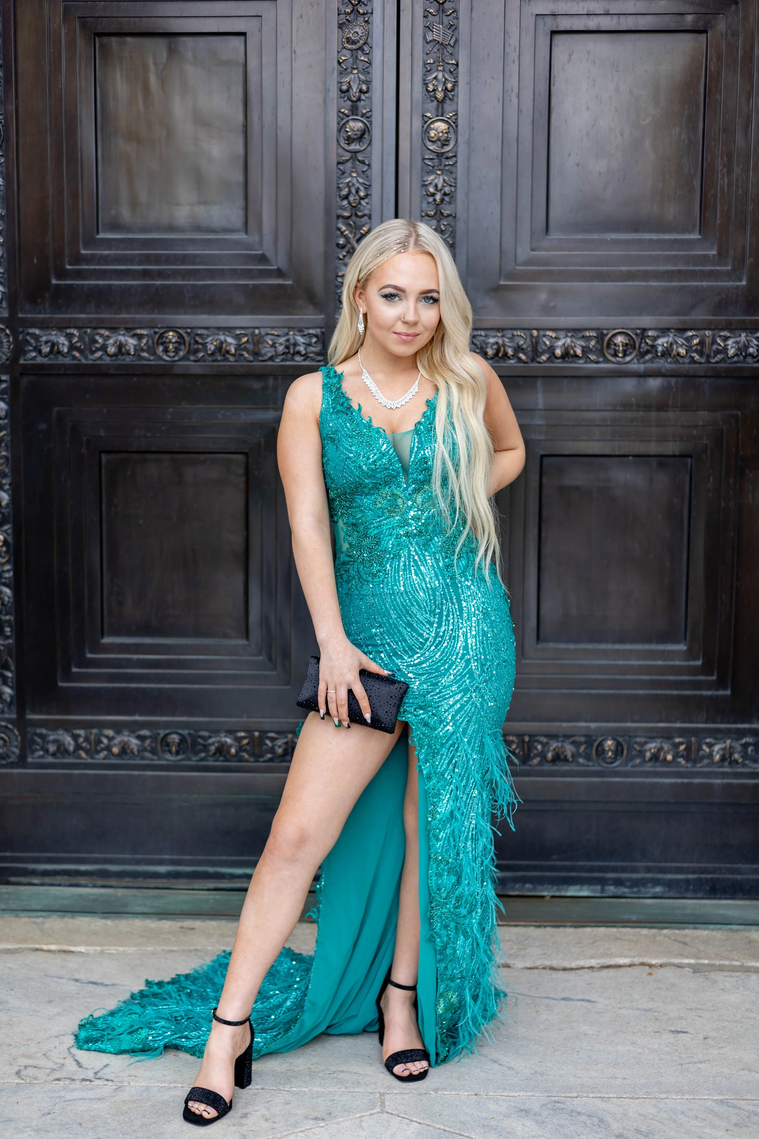 A young woman in an elaborate turquoise gown with intricate beading, a slit revealing her leg, and a train with feathered accents, standing in front of a grand, dark metal door with ornate carvings. She has long blonde hair, wears diamond earrings an