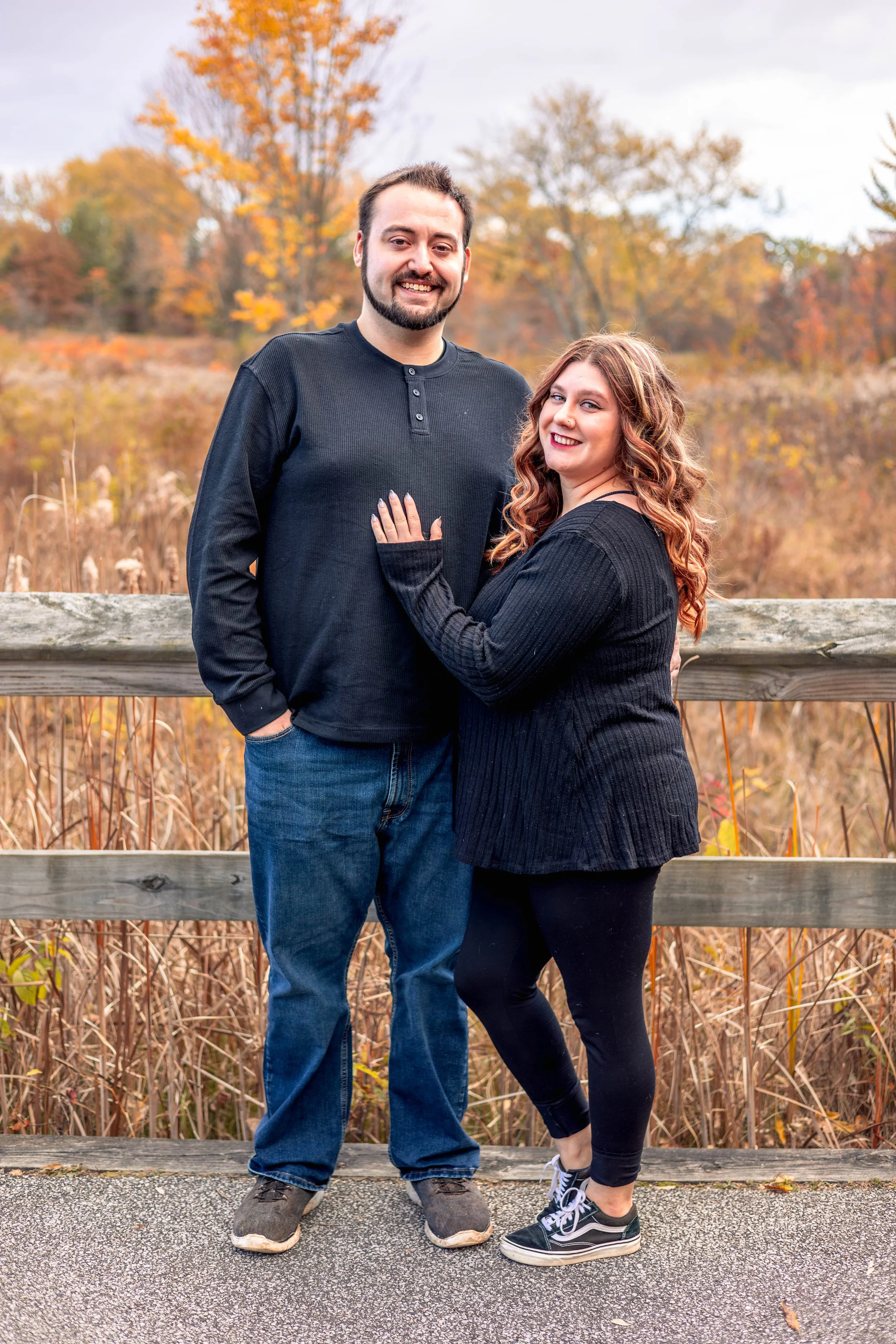 A smiling man and woman standing together outdoors in front of an autumn landscape, with trees showing fall colors, on a wooden bridge railing.