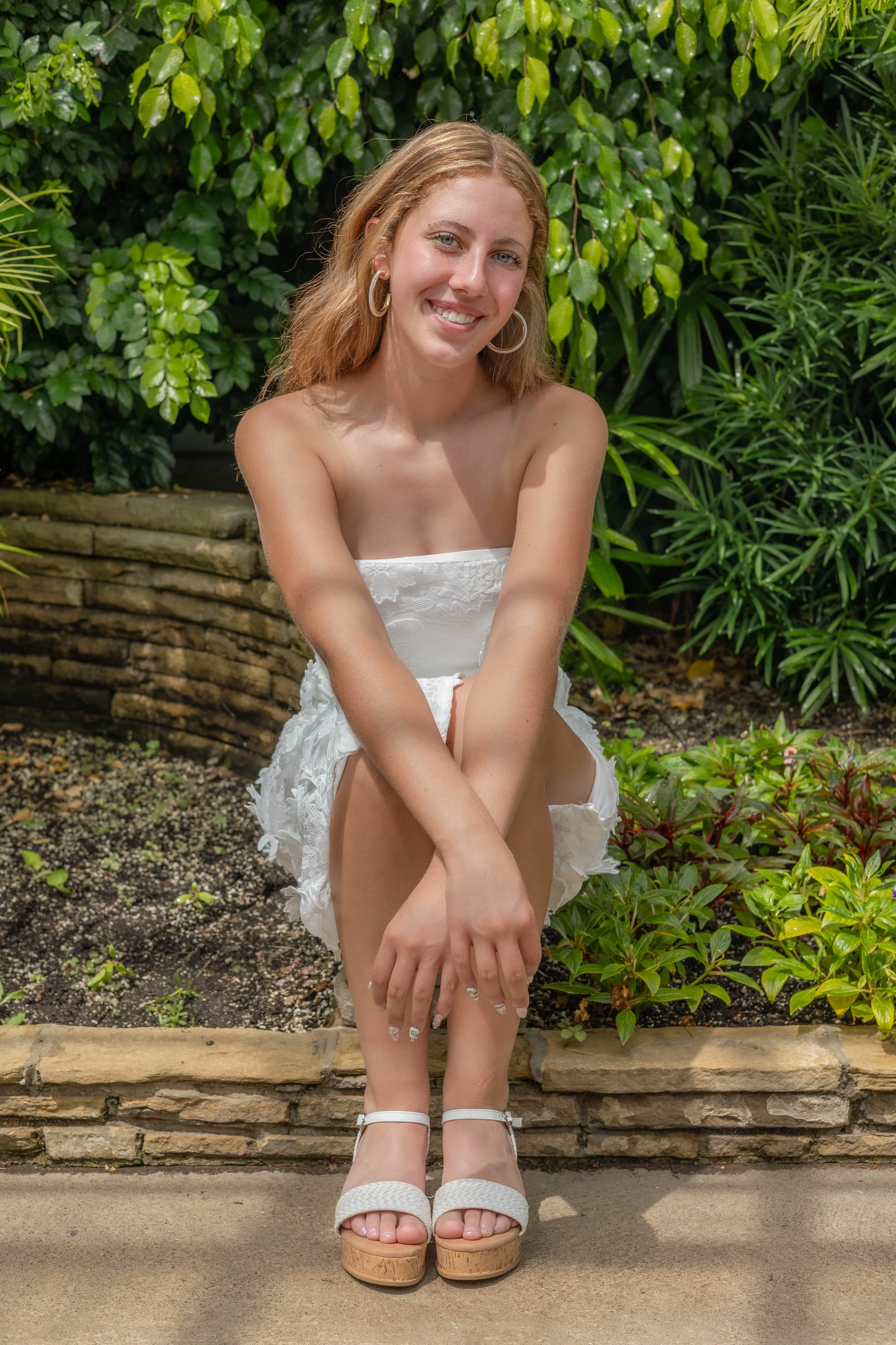 A young woman with red hair and light skin posing outdoors in front of green bushes. She is sitting on a stone ledge, wearing a white strapless dress and white platform sandals. She has hoop earrings and is smiling at the camera with her arms resting on her knees.