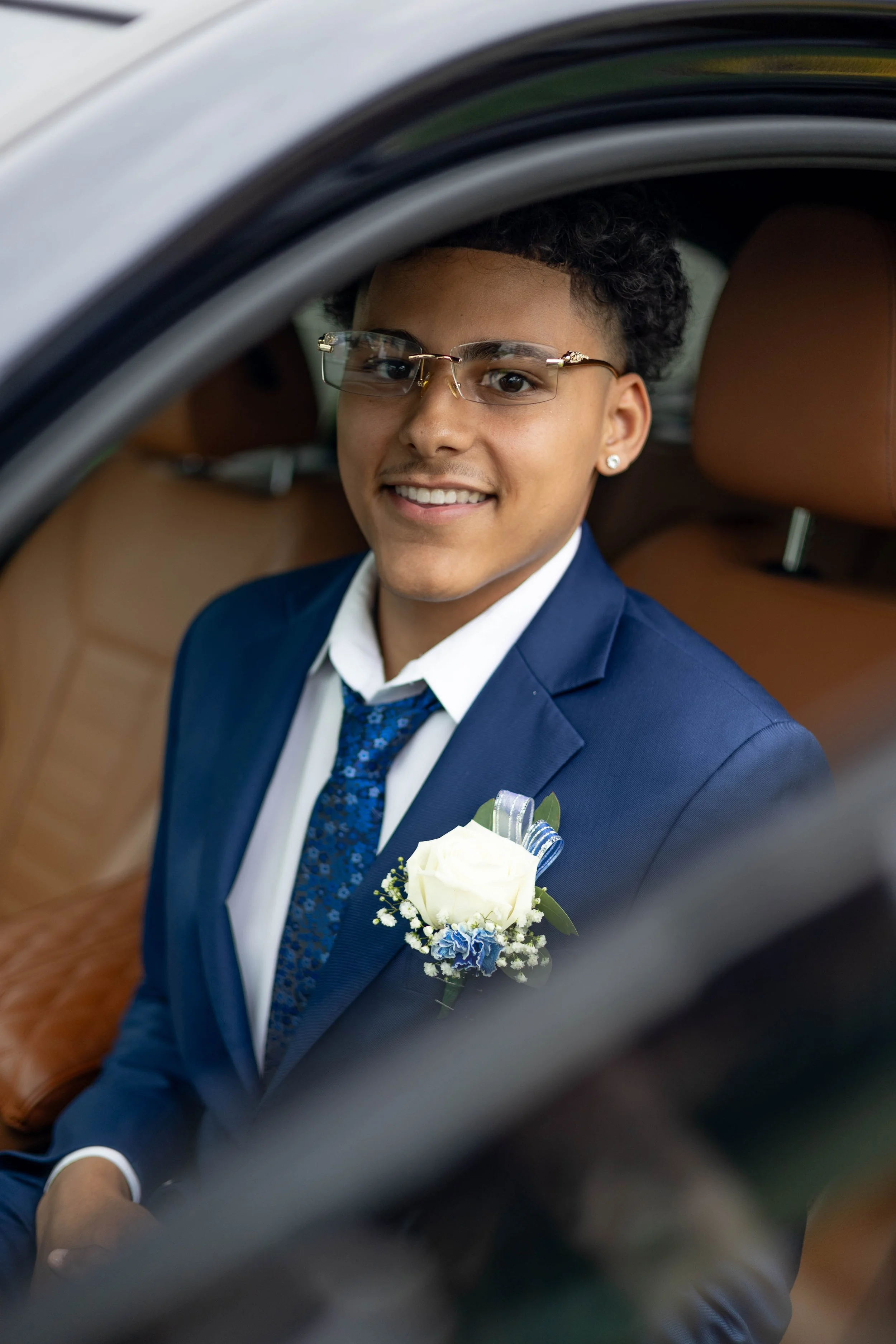 A young man in a blue suit and glasses sitting inside a car, smiling at the camera, with a white flower boutonniere on his suit lapel.
