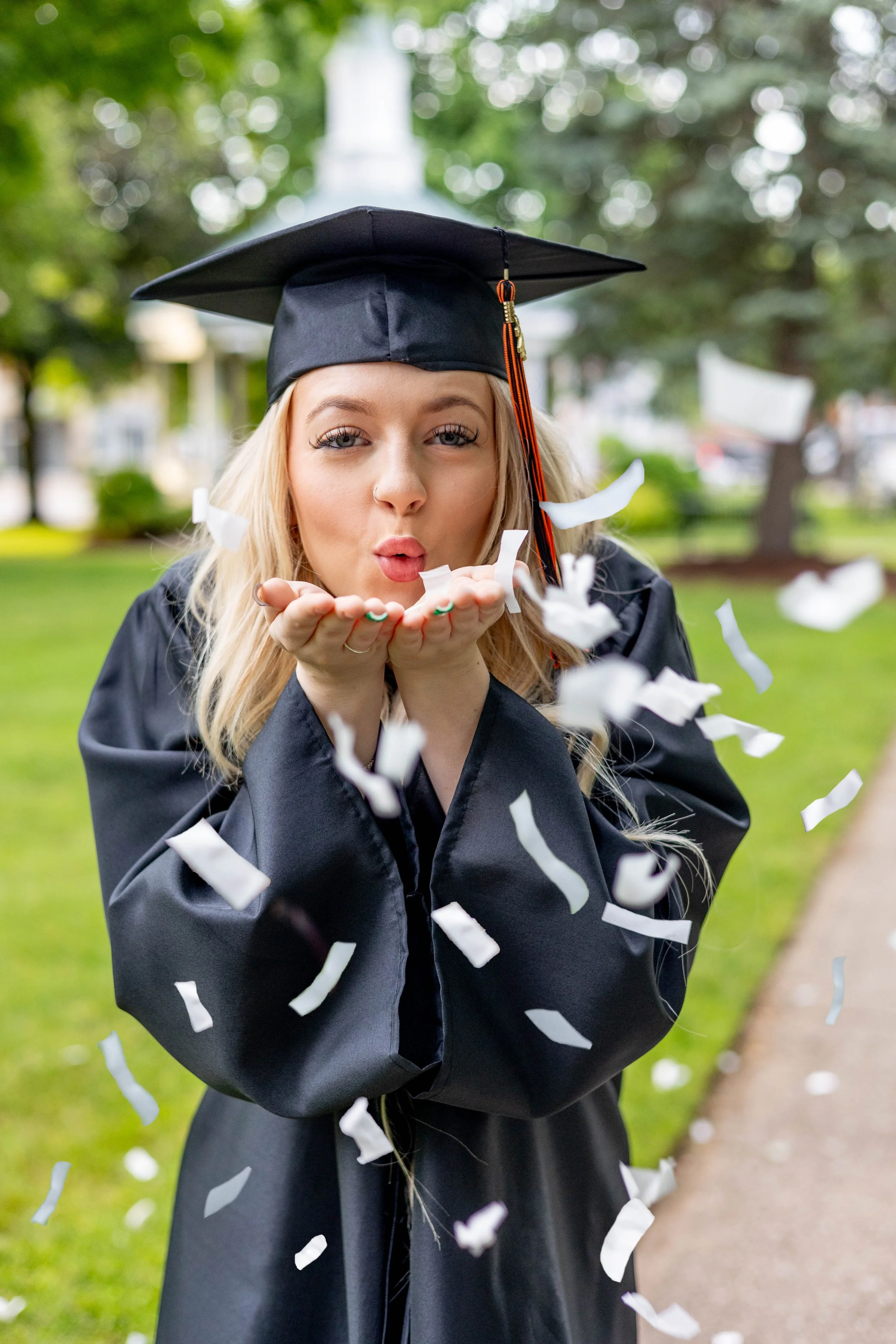 A young woman in a black graduation gown and cap blowing confetti outside in a park with green trees in the background.