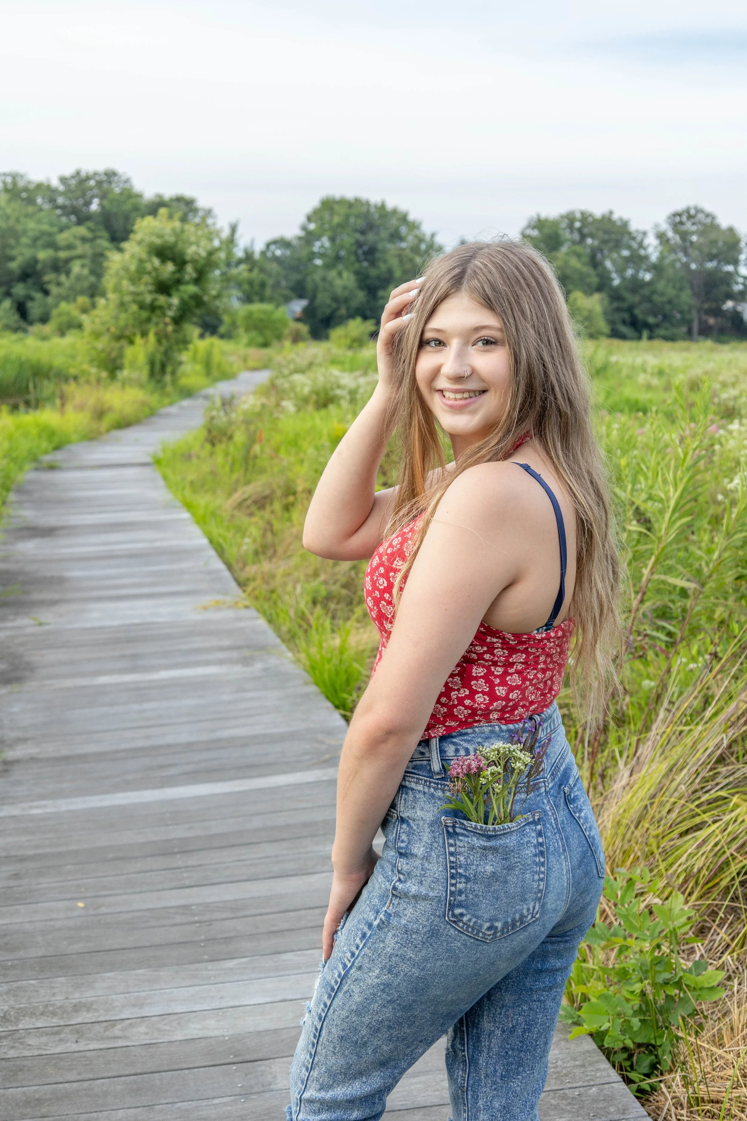 Young woman with long hair, wearing a red floral sleeveless top and blue jeans, smiling, standing on a wooden pathway in a lush green outdoor setting.