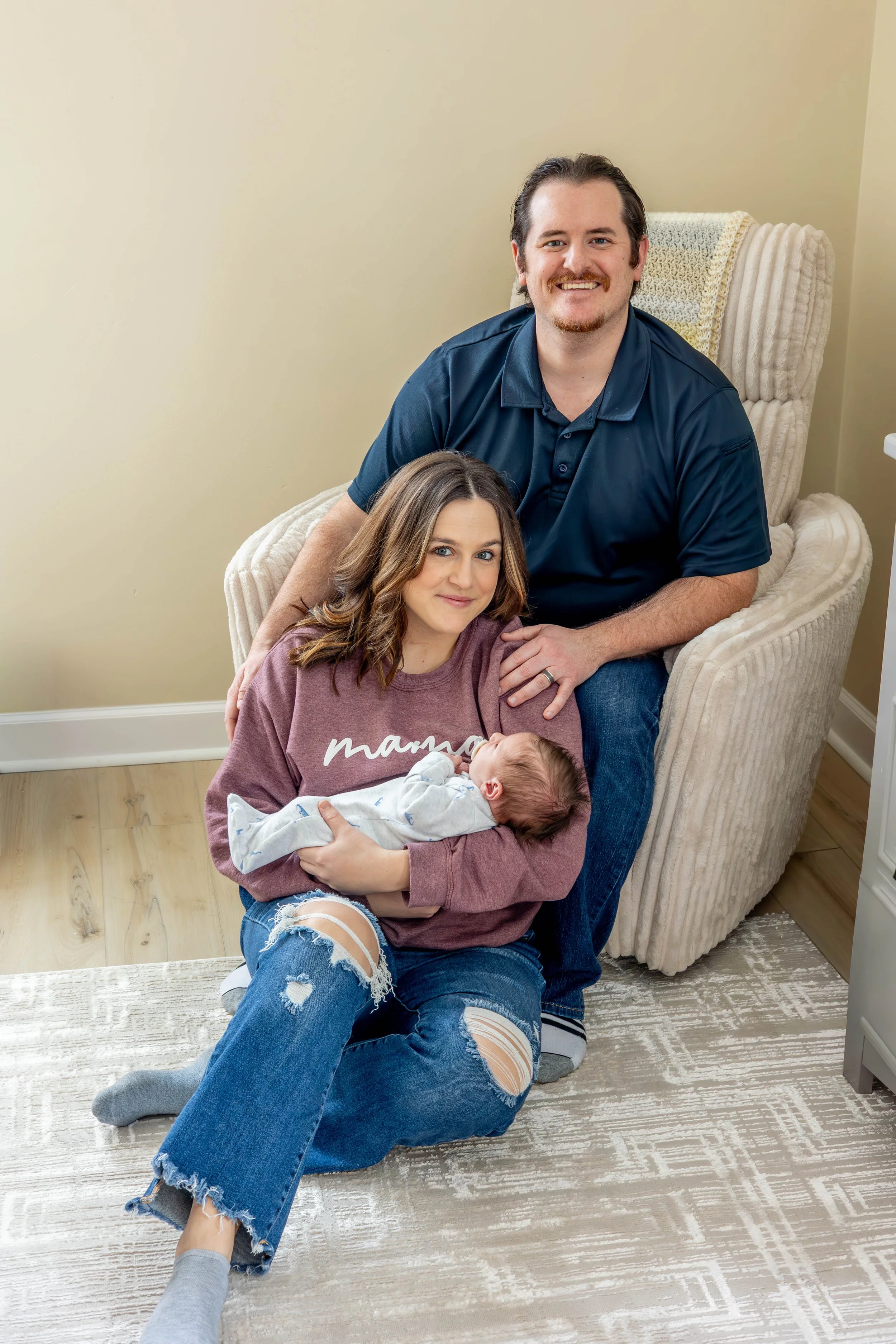 A smiling man sitting in an armchair with a woman holding a newborn baby in her lap, by a woman sitting on the floor in front of them, in a cozy room.
