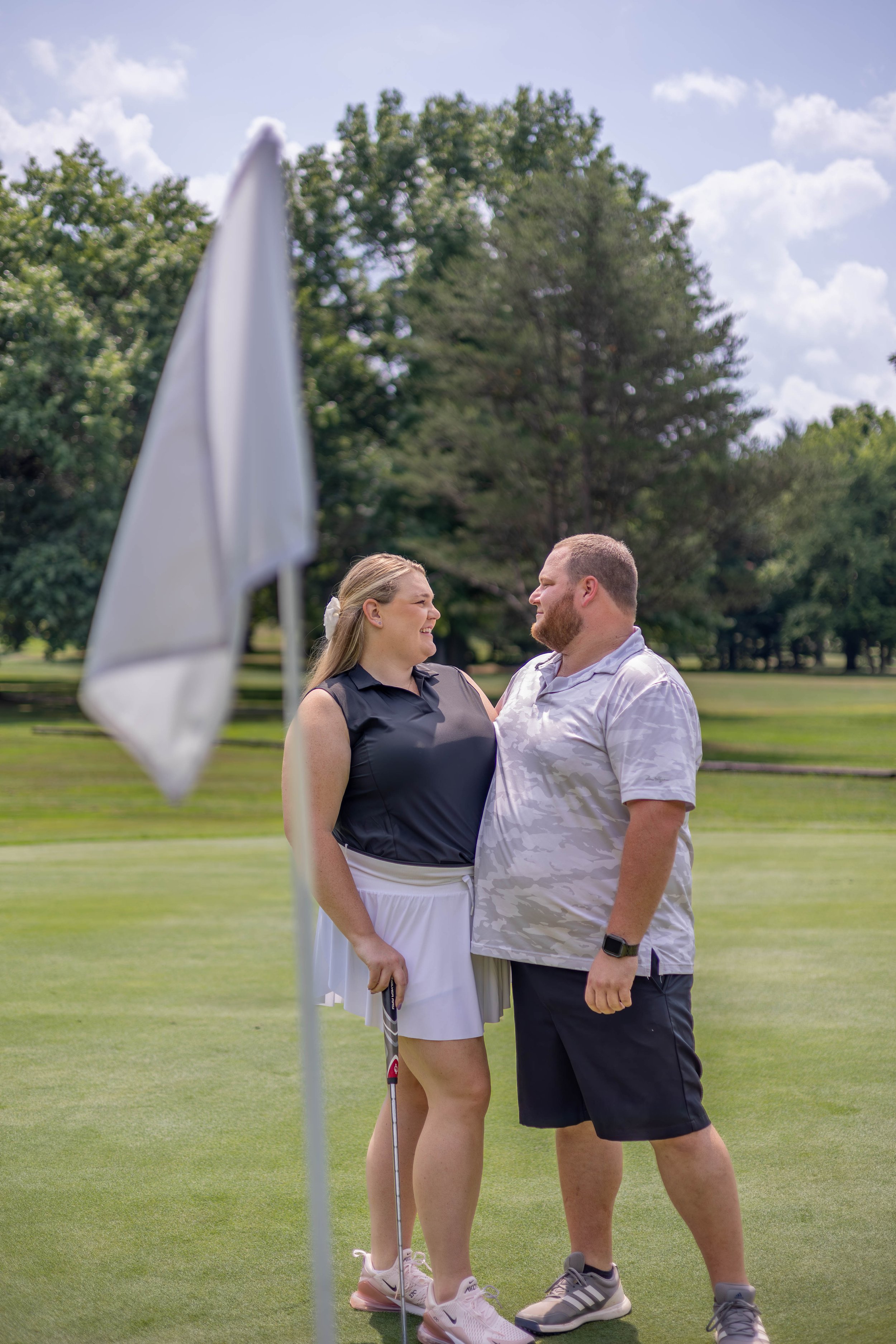 A couple is standing close together on a golf course near a putting green, smiling and looking into each other's eyes. The woman is wearing a sleeveless golf shirt, white skirt, and pink golf shoes, holding a golf club. The man is wearing a white patterned golf shirt, black shorts, and gray shoes, with a golf watch on his wrist. An American flag golf flag is visible in the foreground, and there are trees and a cloudy sky in the background.