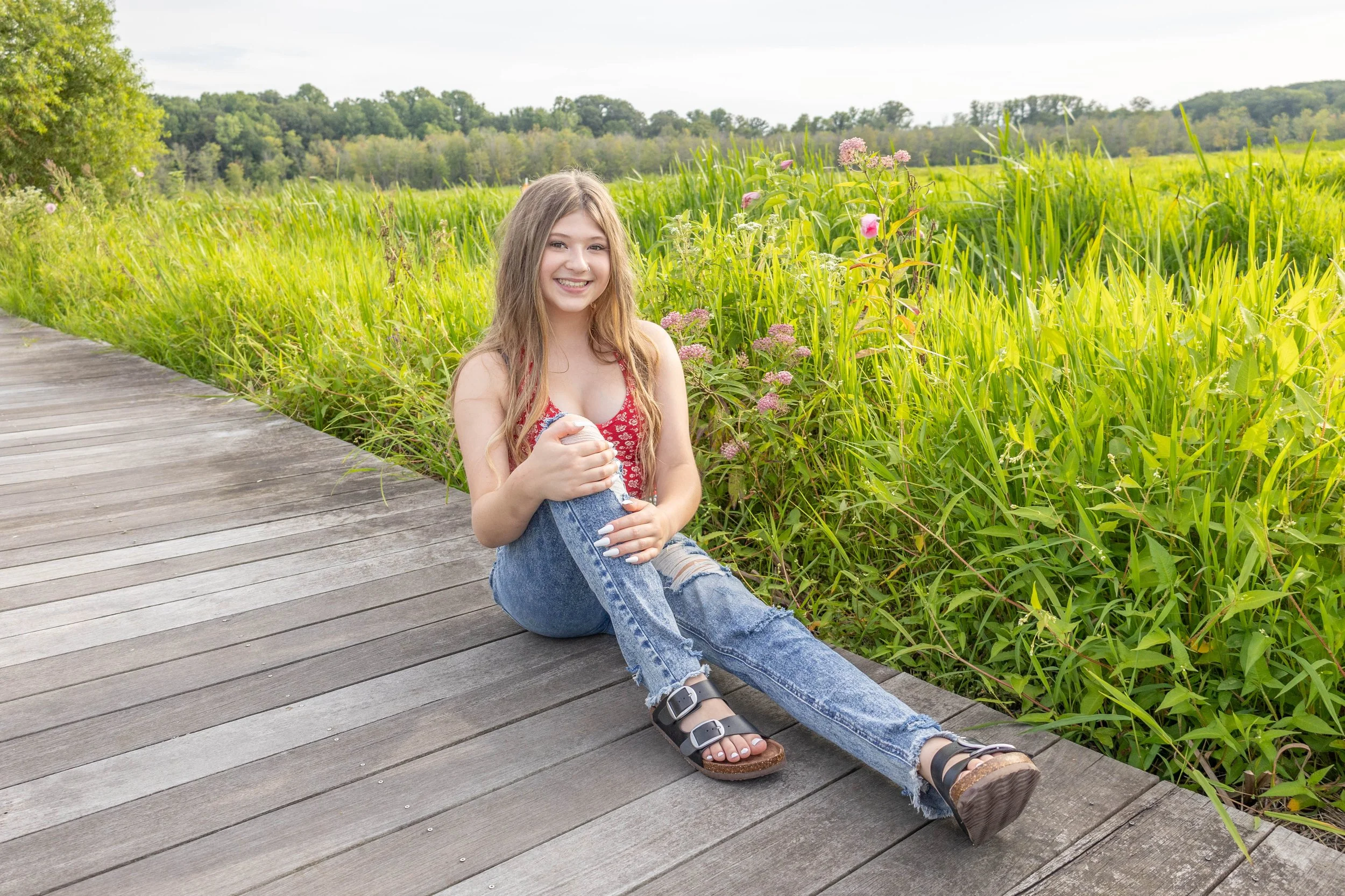 A young girl sits on a wooden pathway beside tall green grass and wildflowers, smiling at the camera in an outdoor setting with trees and hills in the background.