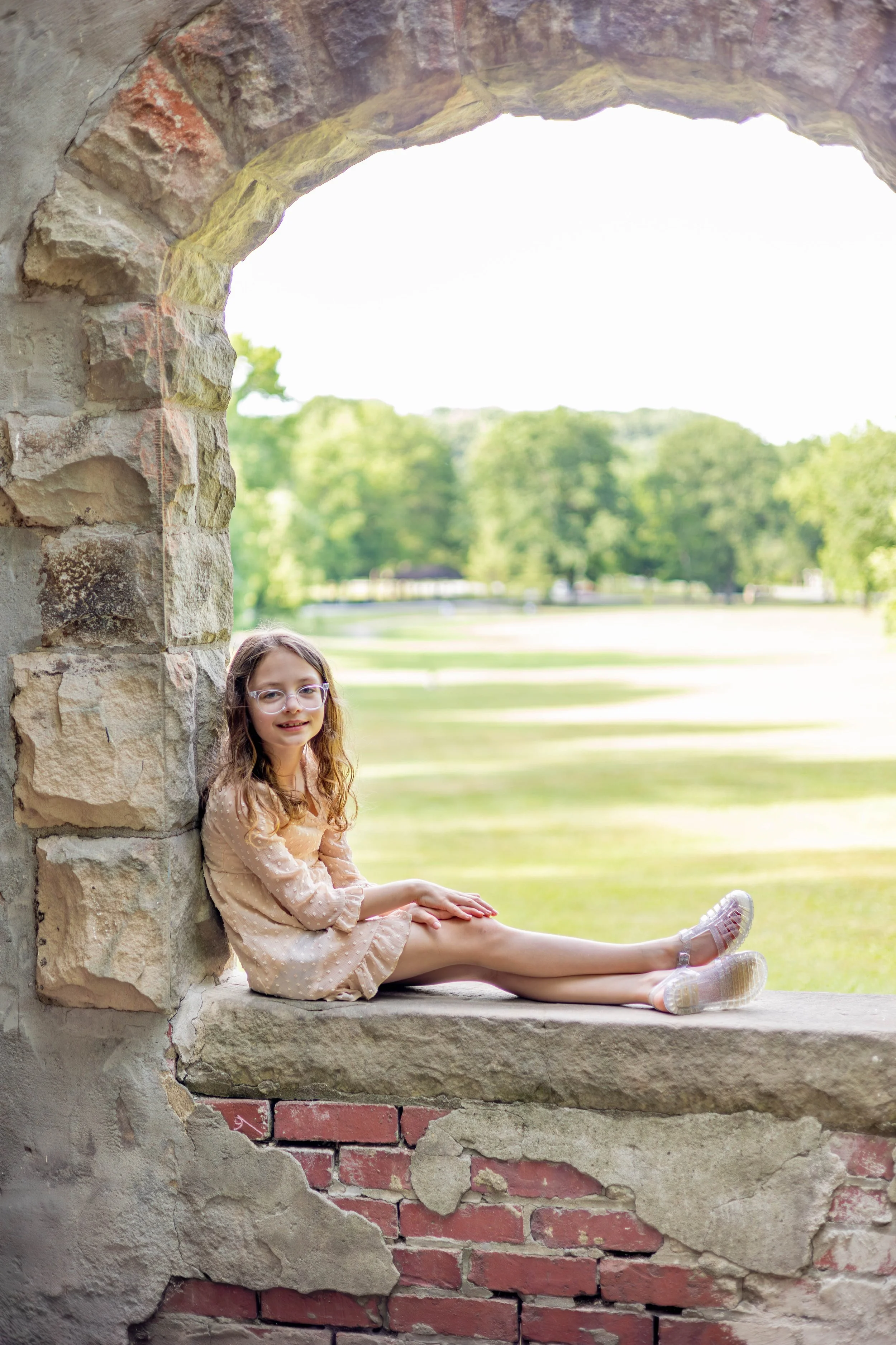 A young girl with glasses and long curly hair sitting on a stone ledge within a brick and stone archway, with a background of green trees and grass.