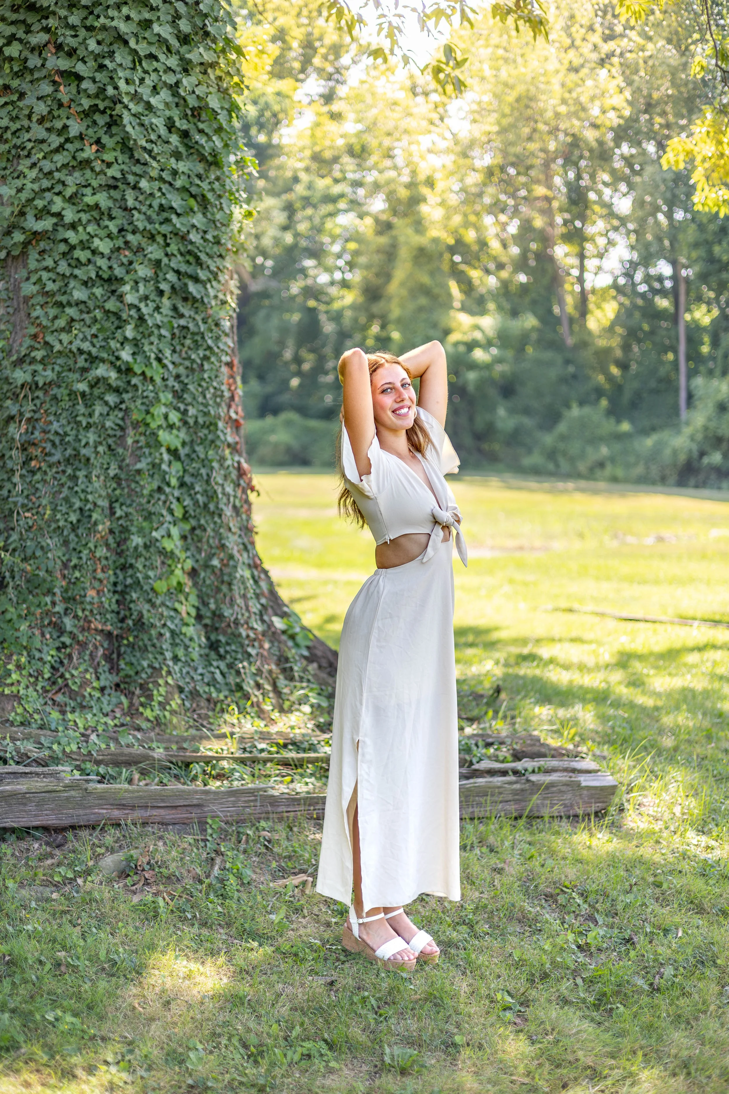 A young woman with long blonde hair, wearing a light-colored crop top and matching long skirt with side slit, standing outdoors near a large ivy-covered tree, smiling with arms behind her head, in a sunlit park setting.