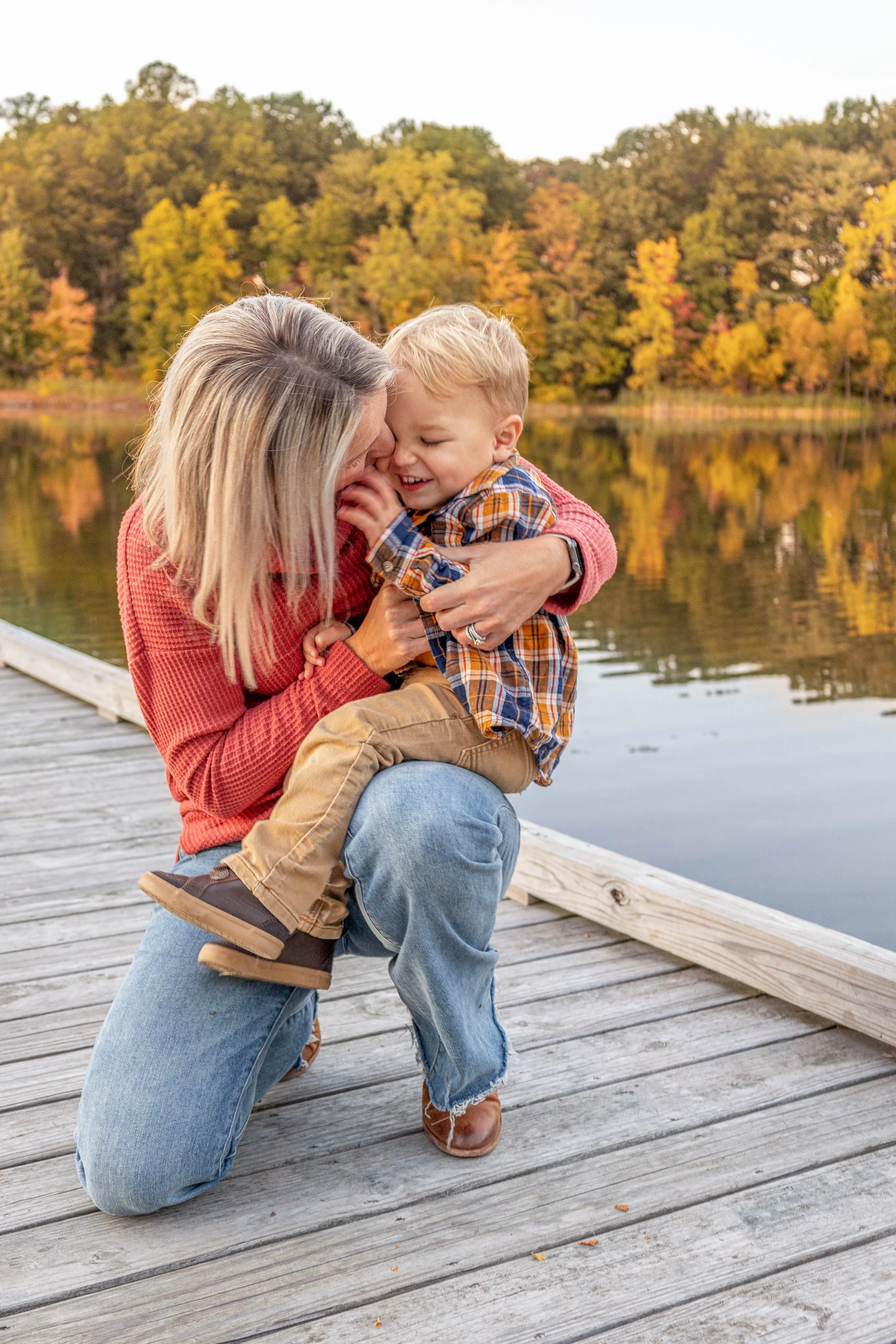 A woman kneeling on a wooden dock, holding and playing with a young boy near a lake with trees displaying fall foliage in the background.