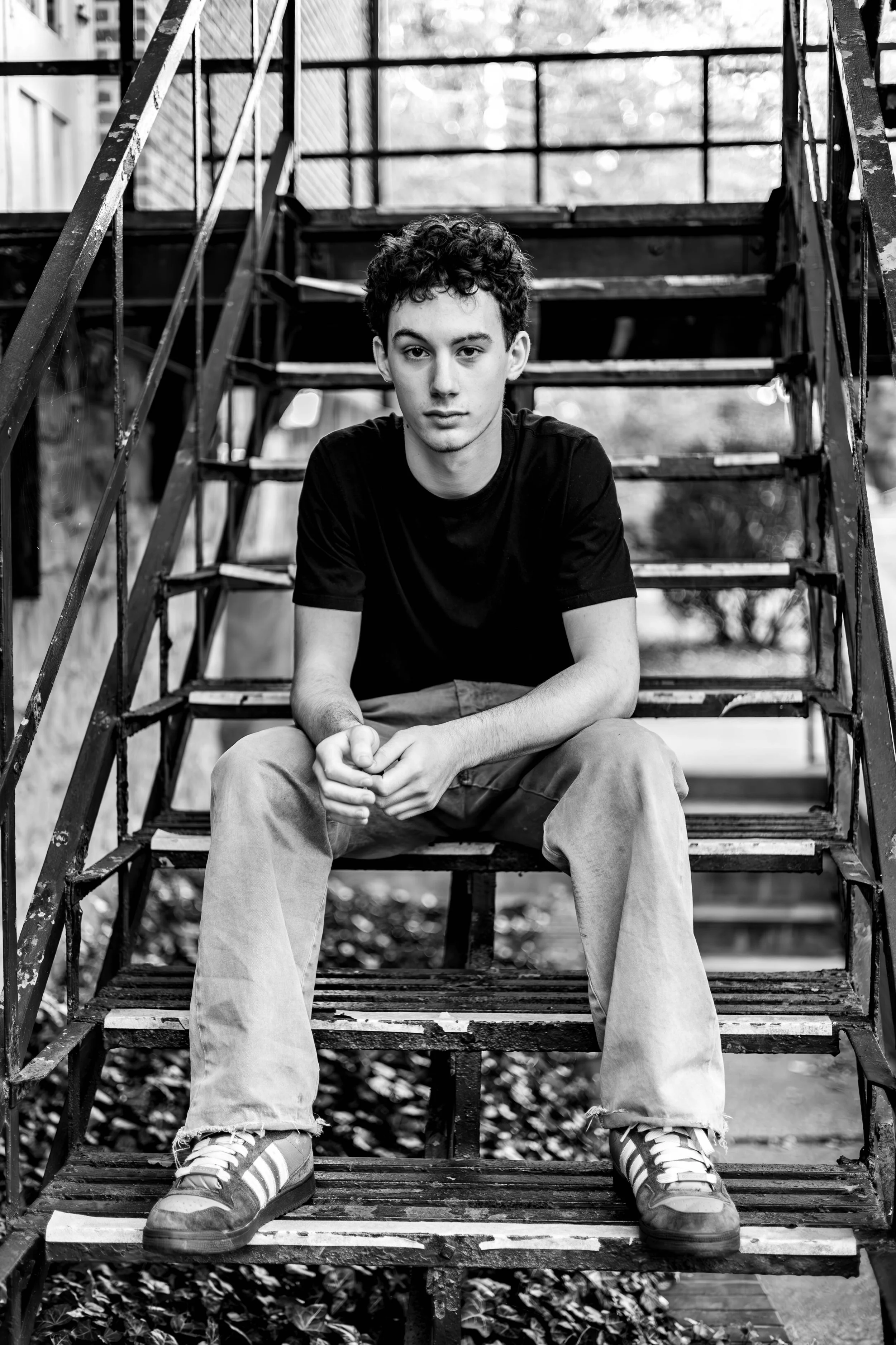 A young man with curly hair sitting on a metal staircase outdoors, looking directly at the camera, in black and white.