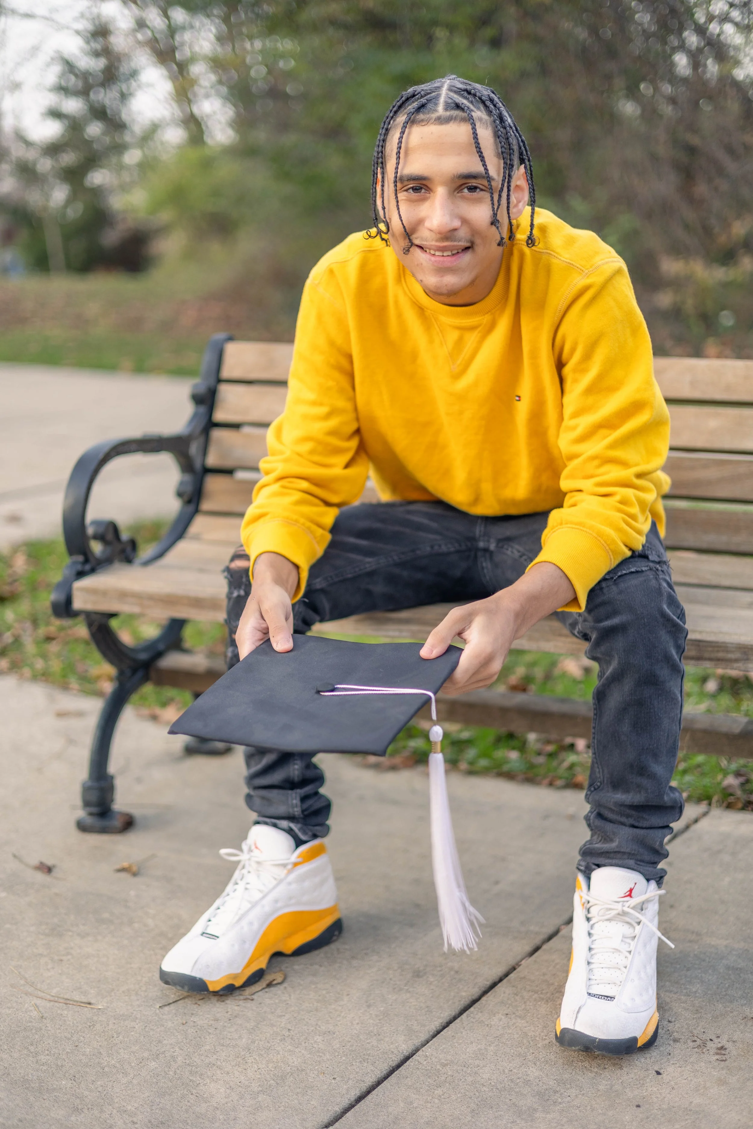 A young man in a yellow sweatshirt and white sneakers is sitting on a park bench, holding a graduation cap with a white tassel, while outdoors with trees in the background.