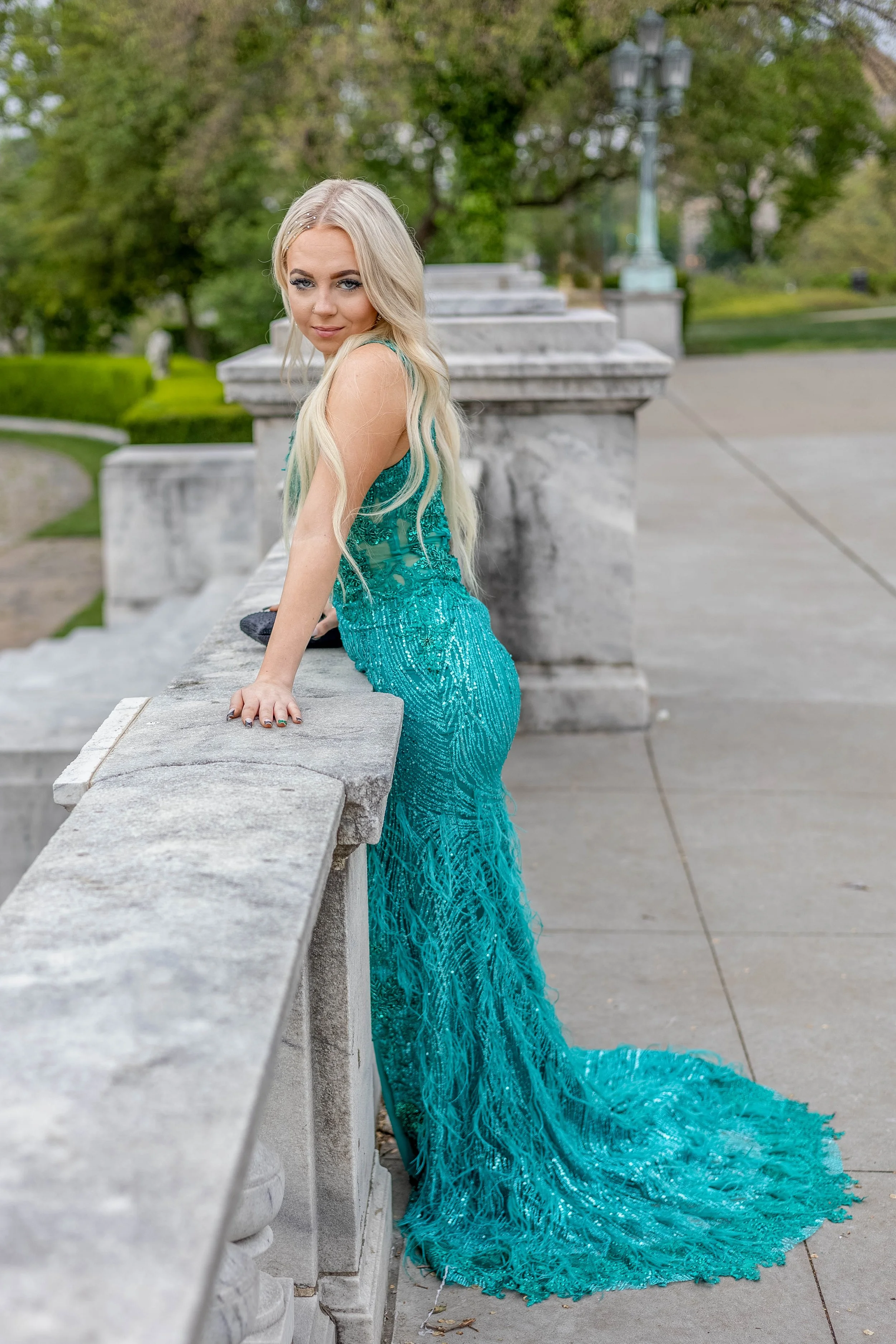 A woman in a turquoise, shimmering gown with lace and fringe detailing, standing outdoors by a stone balustrade, with green trees and a park setting in the background.