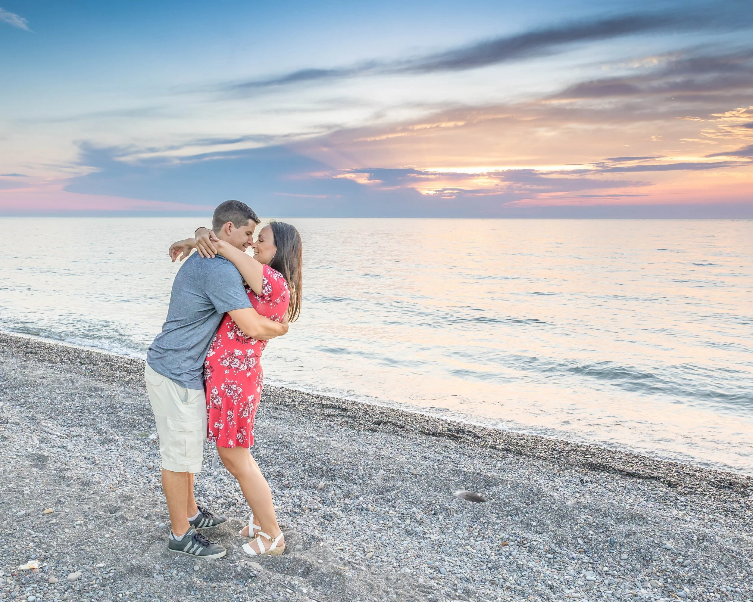 A couple embracing on a pebbled beach during sunset, with the ocean and colorful sky in the background.
