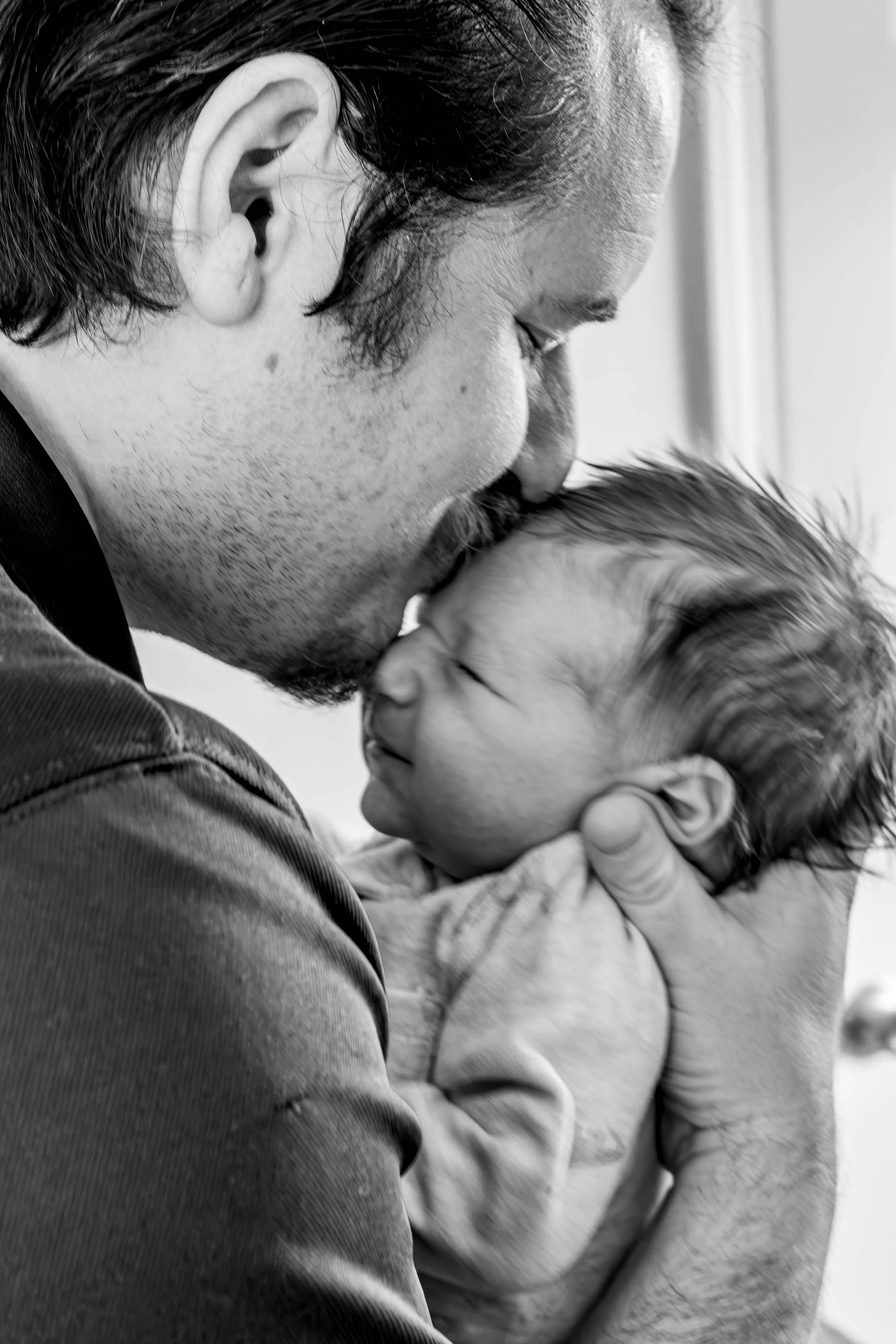 A man with dark hair and stubble is gently kissing a young child's head. The child has light hair and is wearing a light-colored jacket. The image is in black and white.