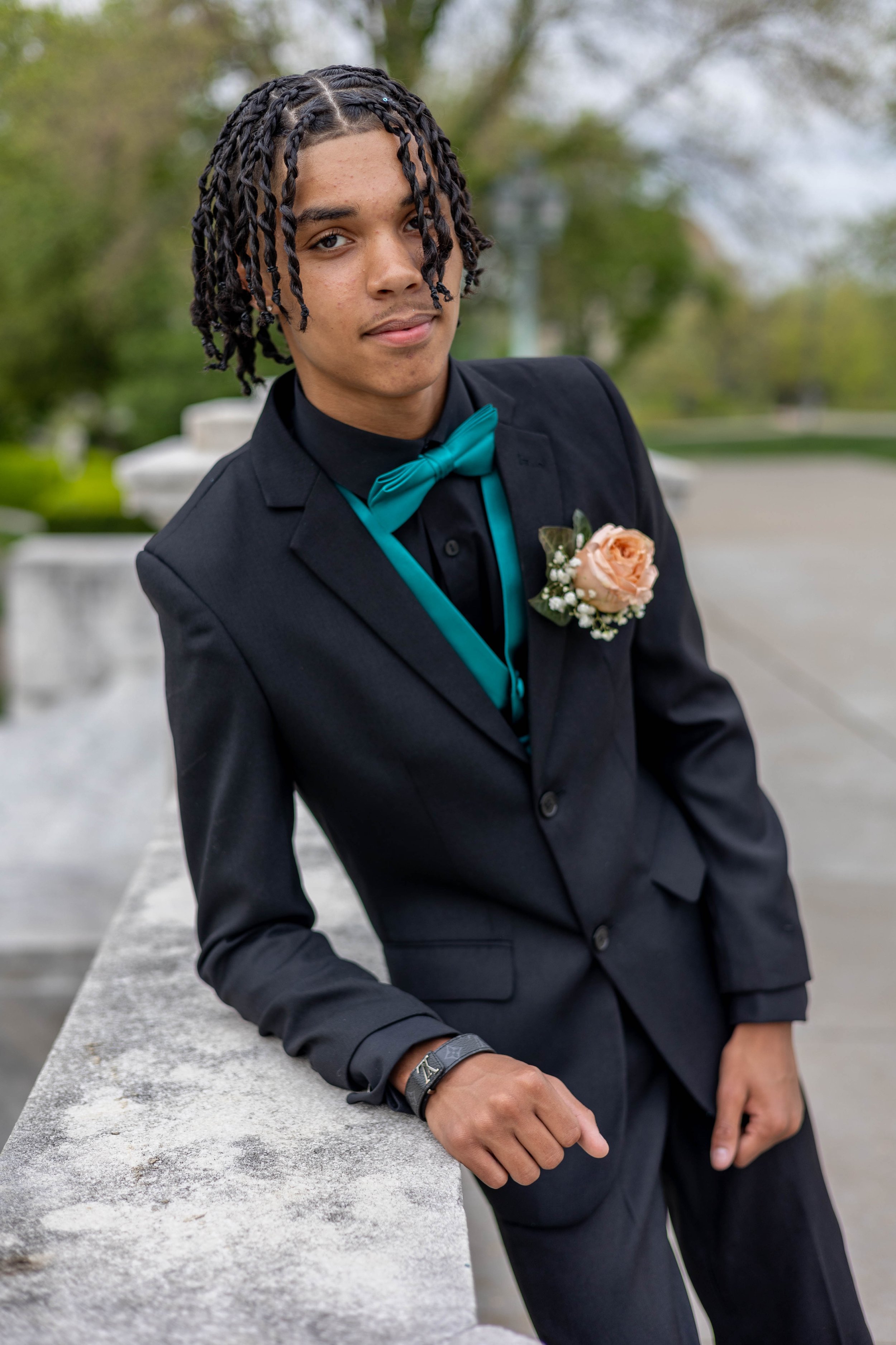 Young man in a black suit with a teal bow tie and boutonniere, leaning on a stone ledge outdoors.