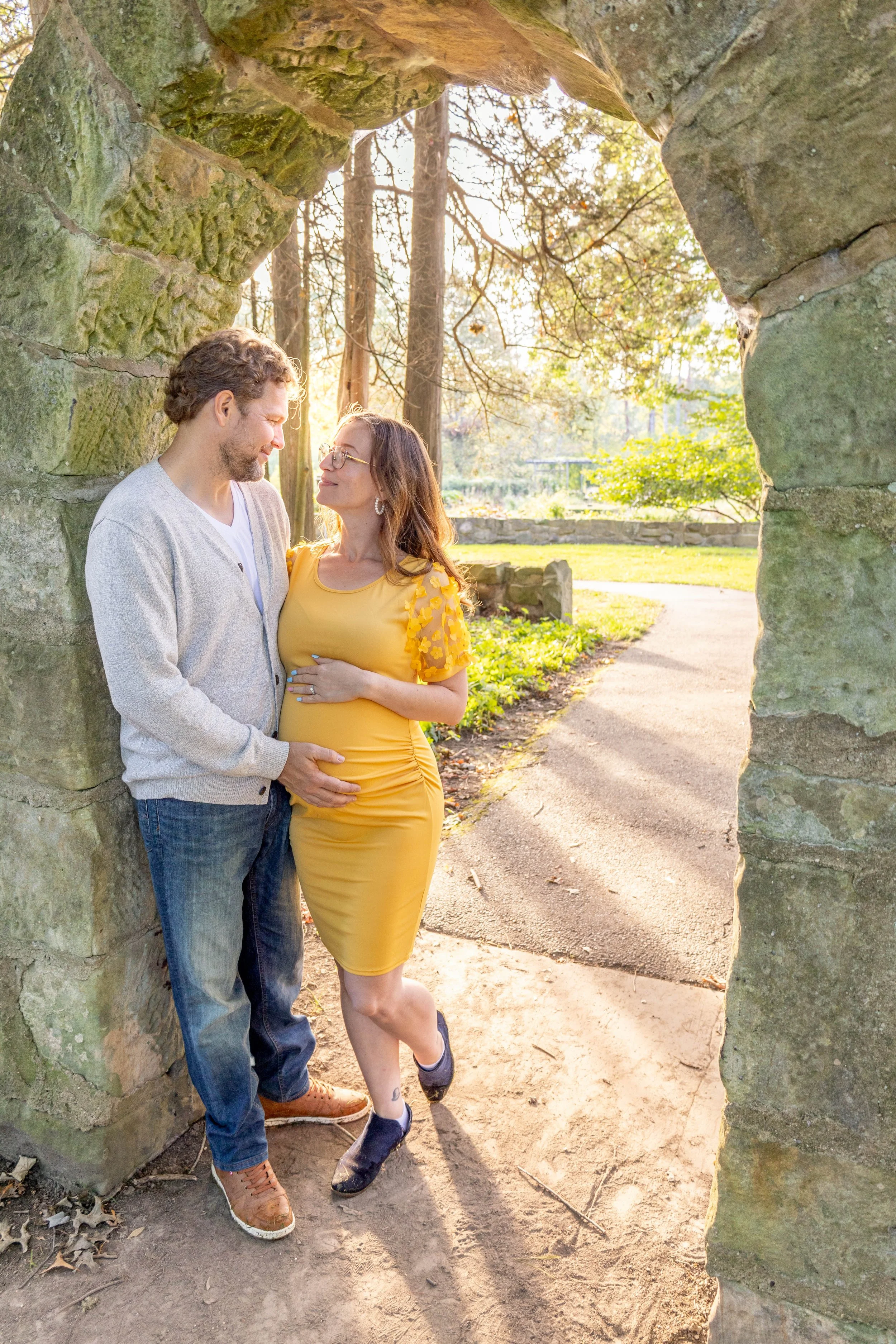 A couple standing close together outdoors near a stone archway, with their heads touching and hands on each other's stomachs, in a park-like setting during late afternoon or early evening.