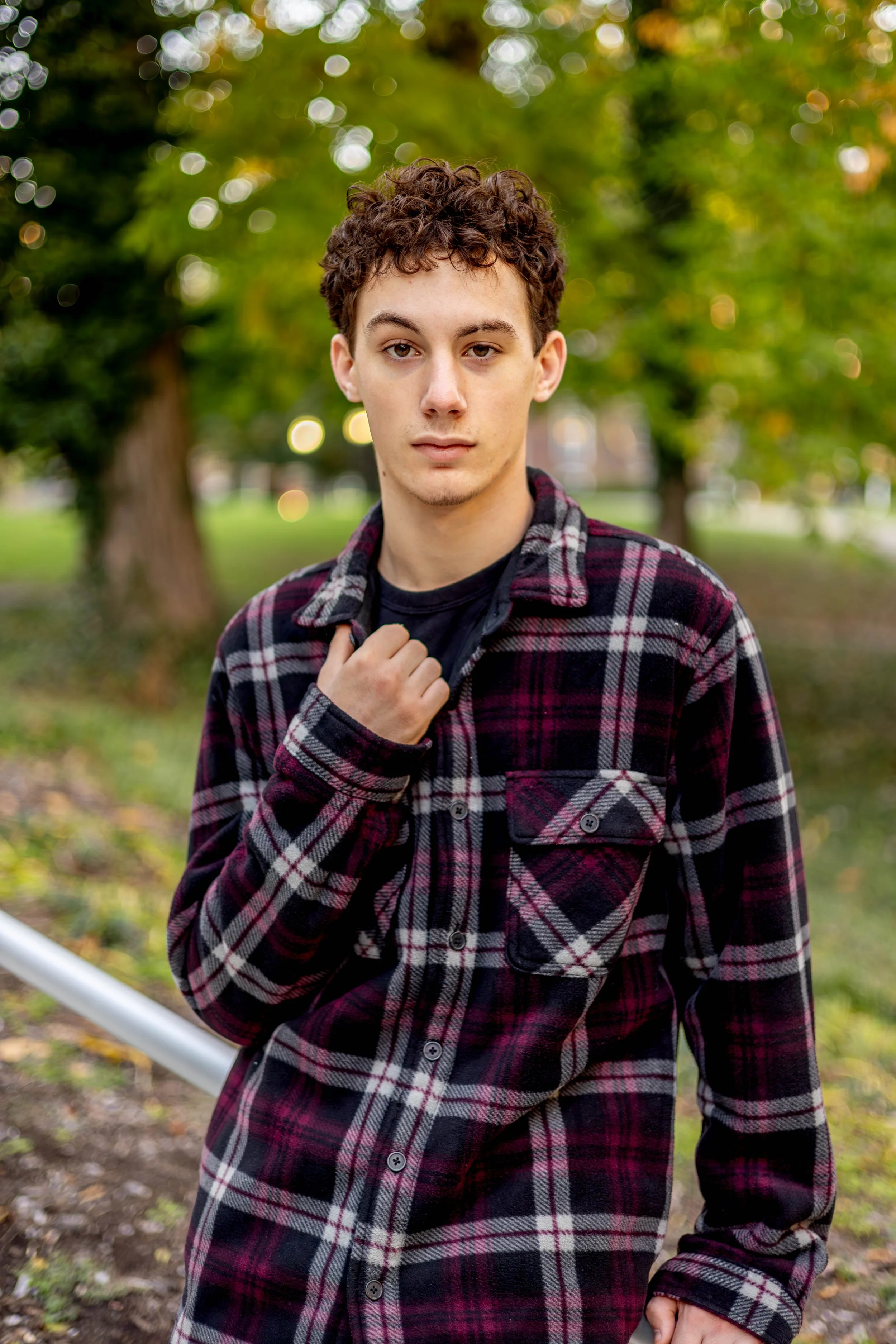 A young man with curly brown hair wearing a black shirt and a red, black, and white plaid jacket standing outdoors in a park with green trees and blurred lights in the background.