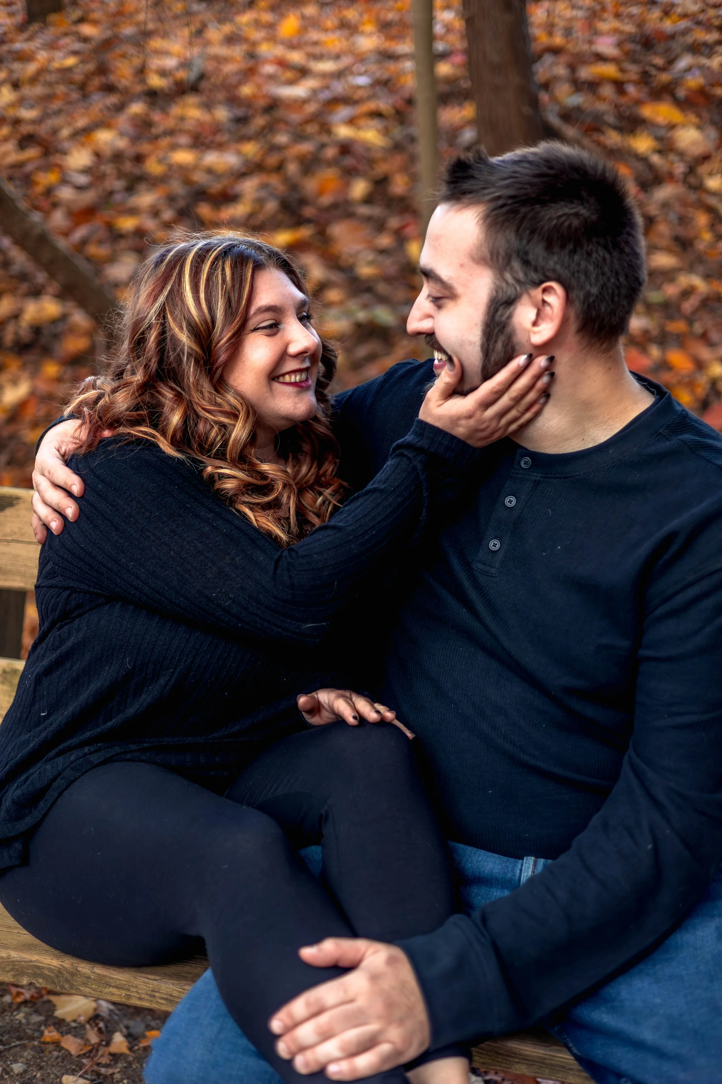 A couple sitting on a wooden bench outdoors in an autumn setting, smiling at each other, with fall leaves on the ground and trees in the background.