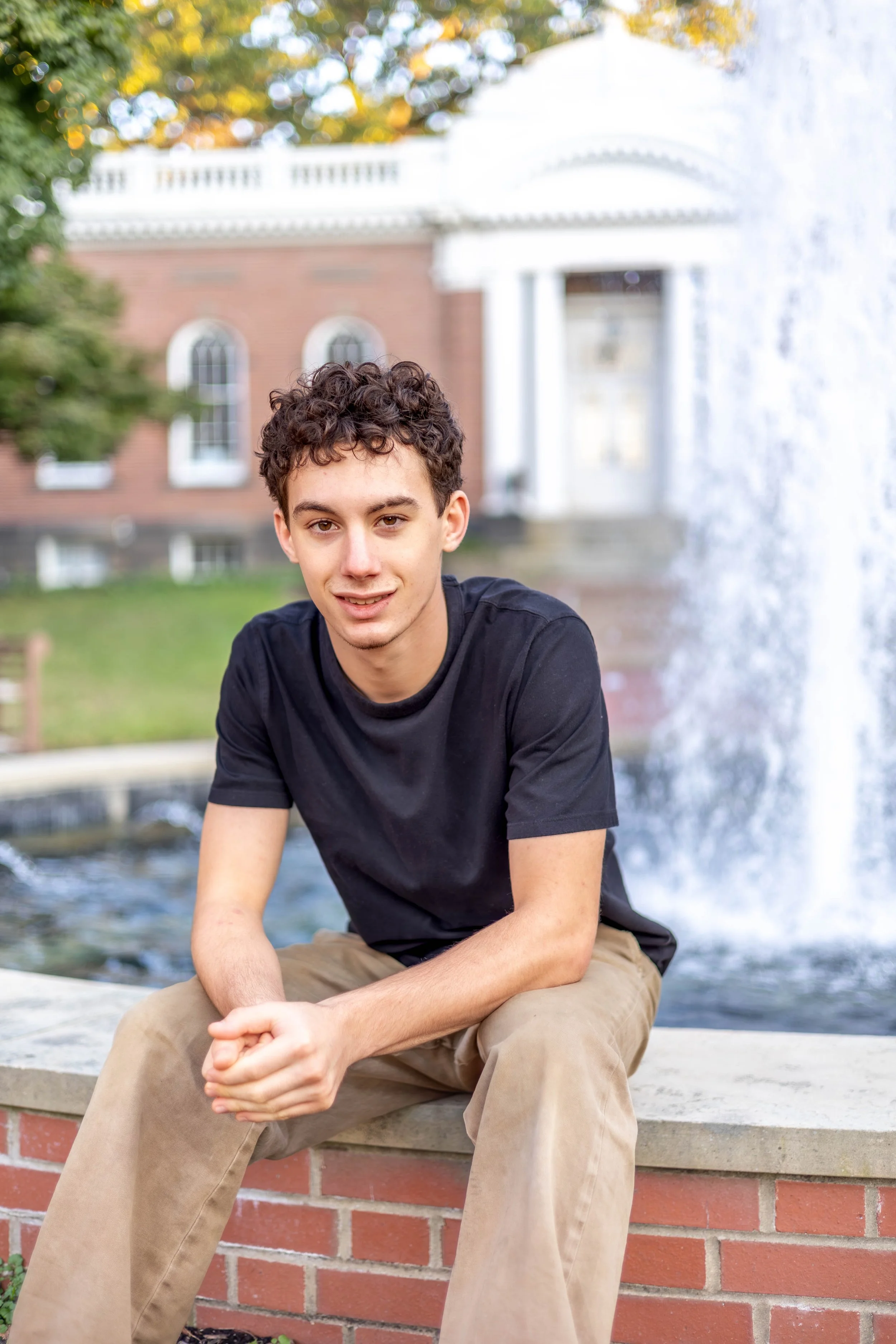 A young man with curly brown hair sitting on a brick ledge near a fountain. He is wearing a black T-shirt and beige pants, smiling slightly. Behind him, there is a large building with arched windows and a white columned entrance, surrounded by trees with autumn-colored leaves.