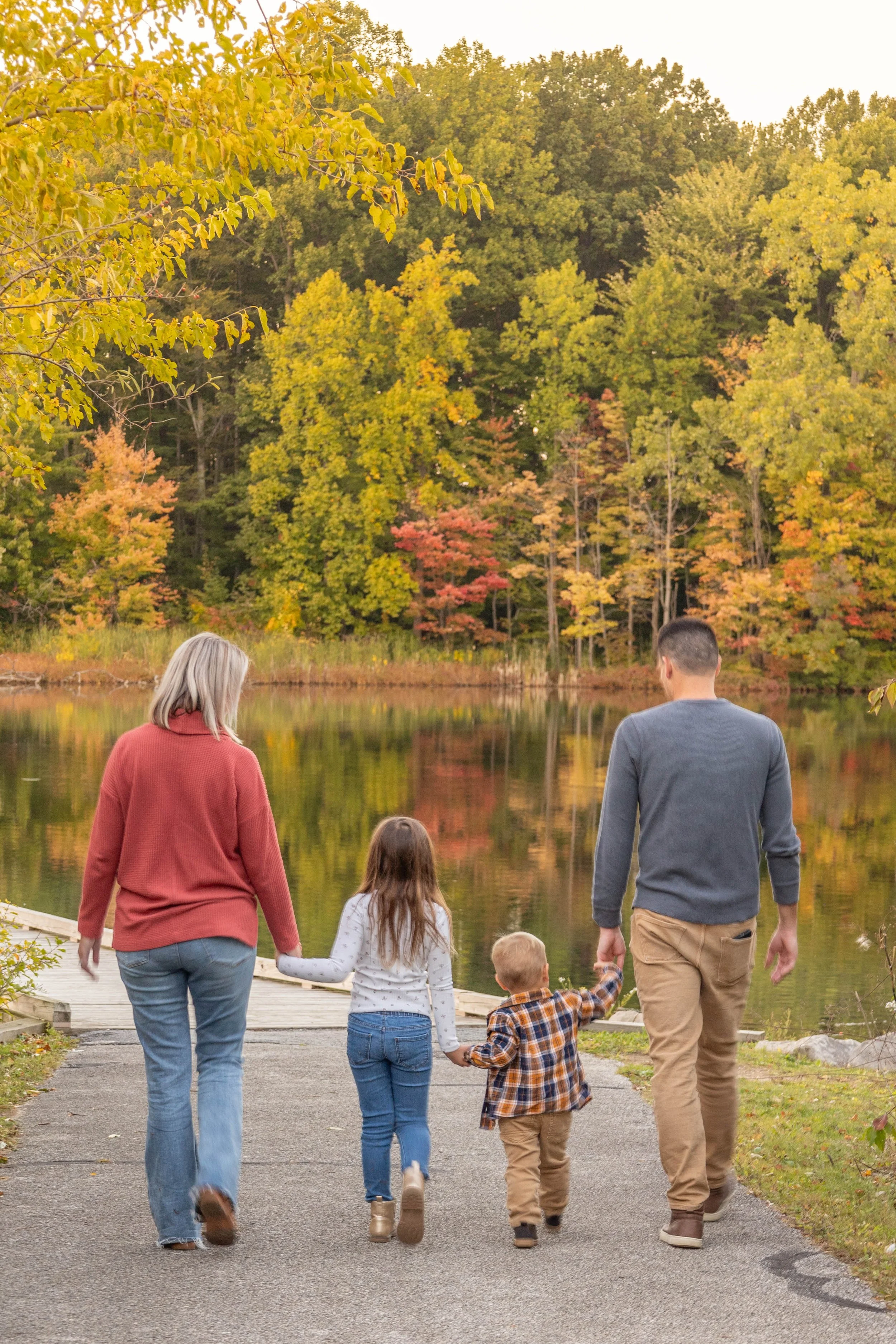 A family walking hand in hand along a paved path by a lake in a park with colorful autumn leaves and trees in the background.