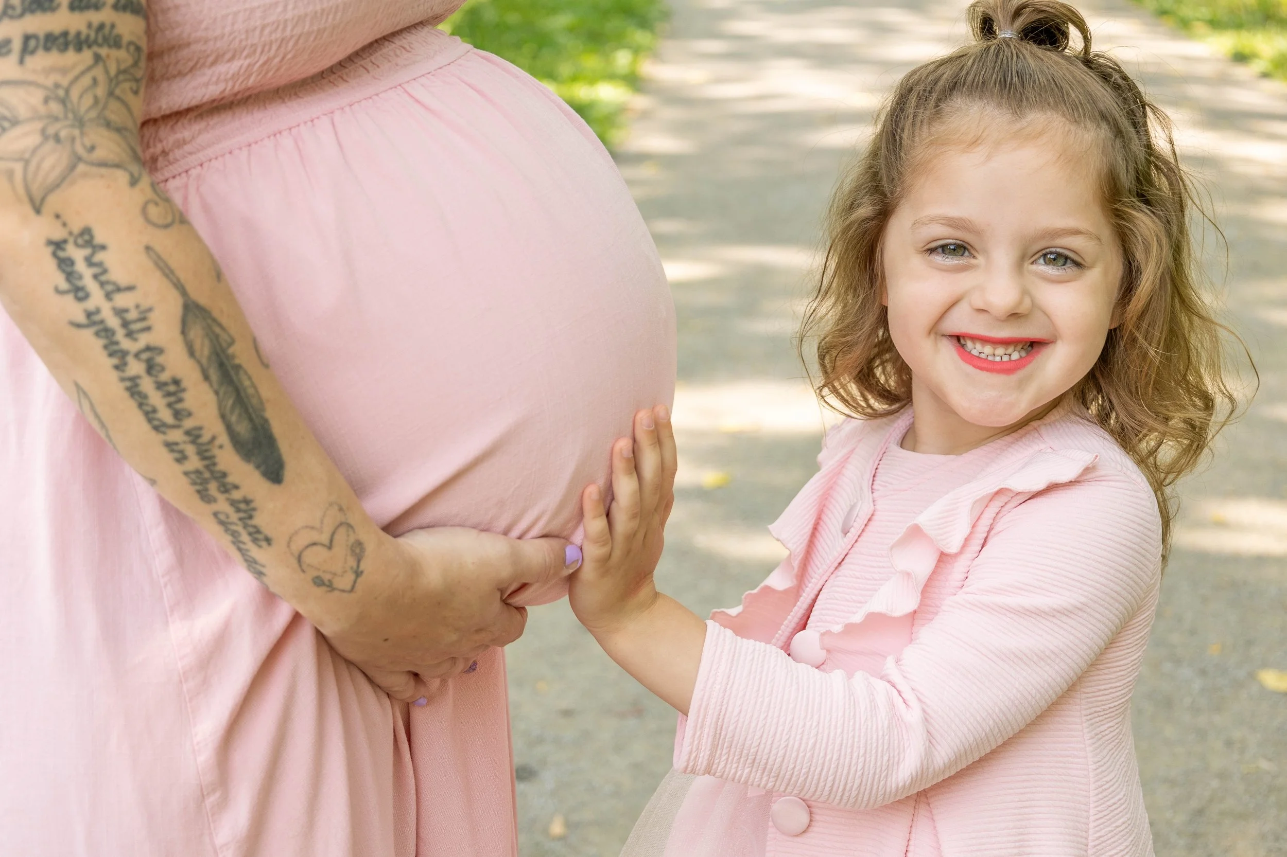 A smiling young girl with curly hair and a pink shirt touches a pregnant woman's belly, who is wearing a pink dress, in an outdoor setting with trees and a pathway.
