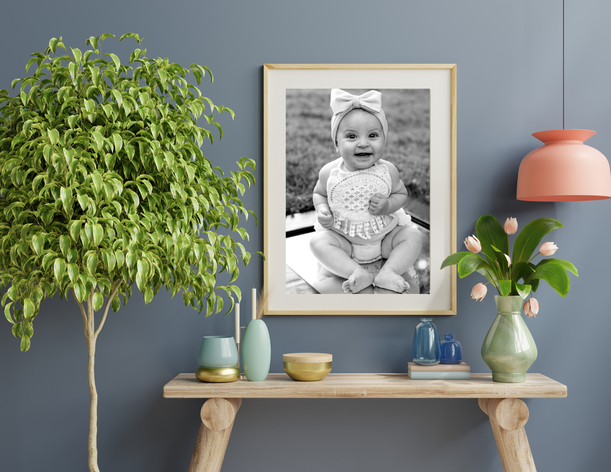 A framed black and white photo of a smiling baby girl with a headband and a lacy dress, surrounded by colorful decorative vases and a green leafy plant on a wooden table against a blue wall.