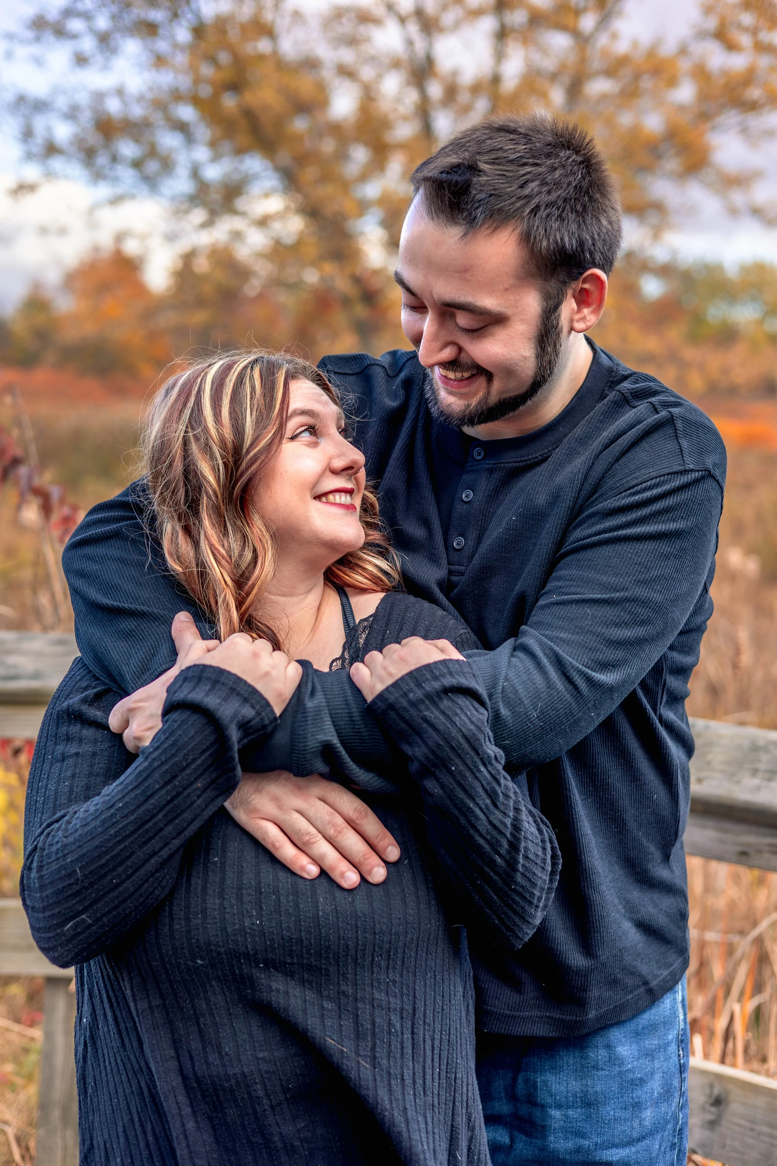 A man and woman smiling and embracing outdoors during autumn, with colorful fall trees in the background.
