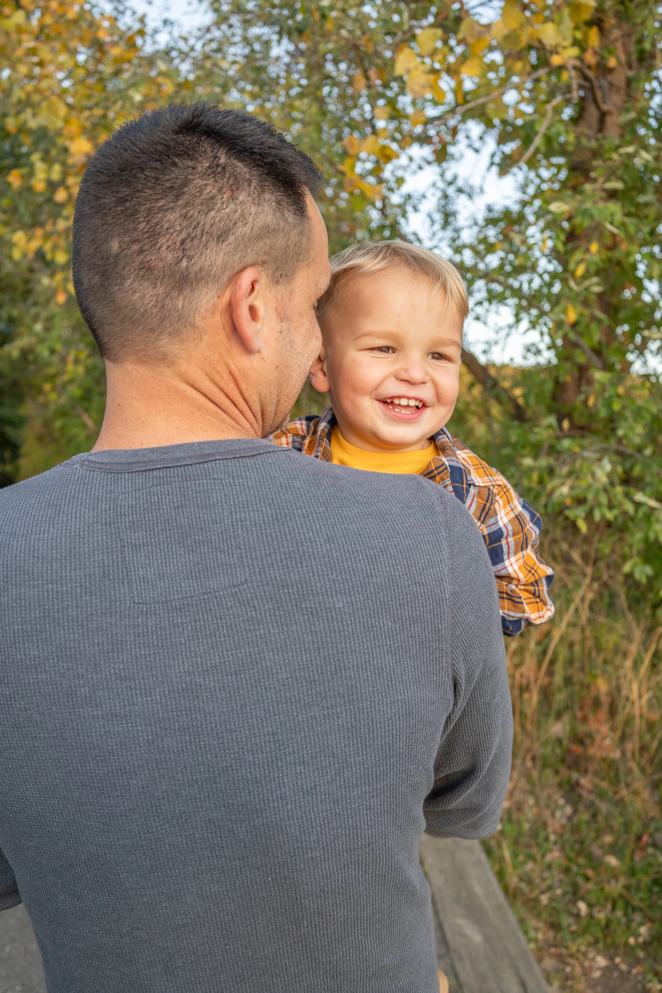 A man holding a young boy outdoors during fall, with trees and foliage in the background.