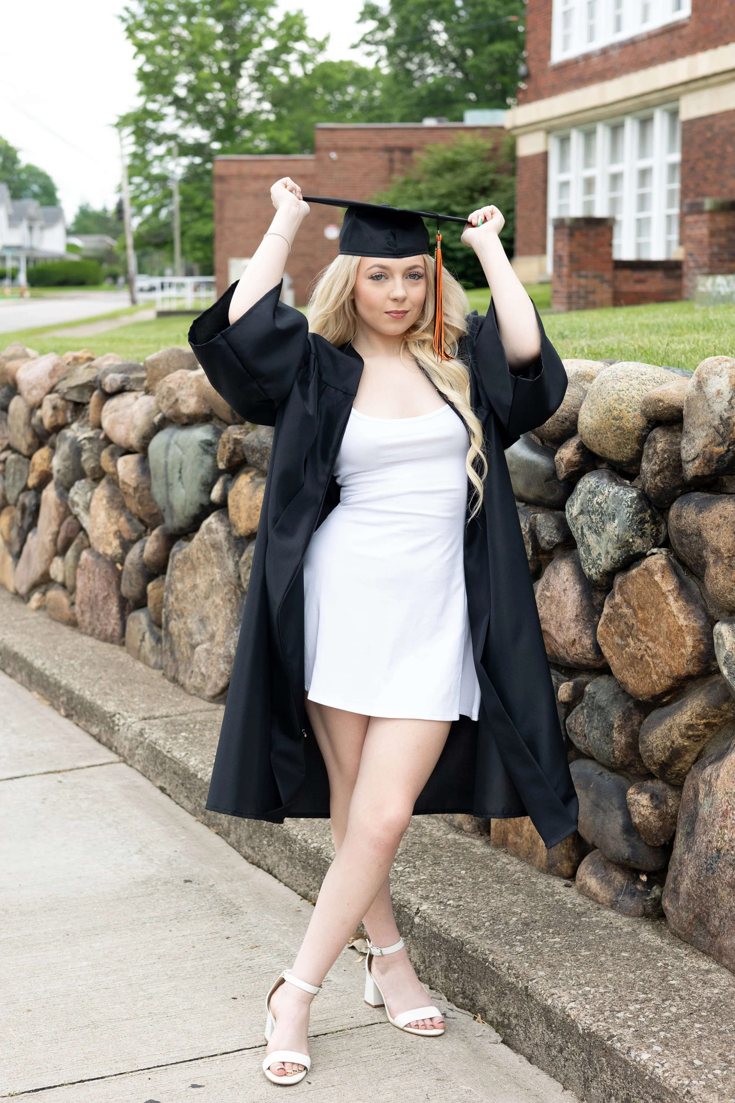 A young woman in a graduation gown and cap standing outdoors near a stone wall. She is adjusting her cap with both hands, wearing white high-heeled shoes, and looking at the camera with a confident expression. Green trees and brick buildings are in t