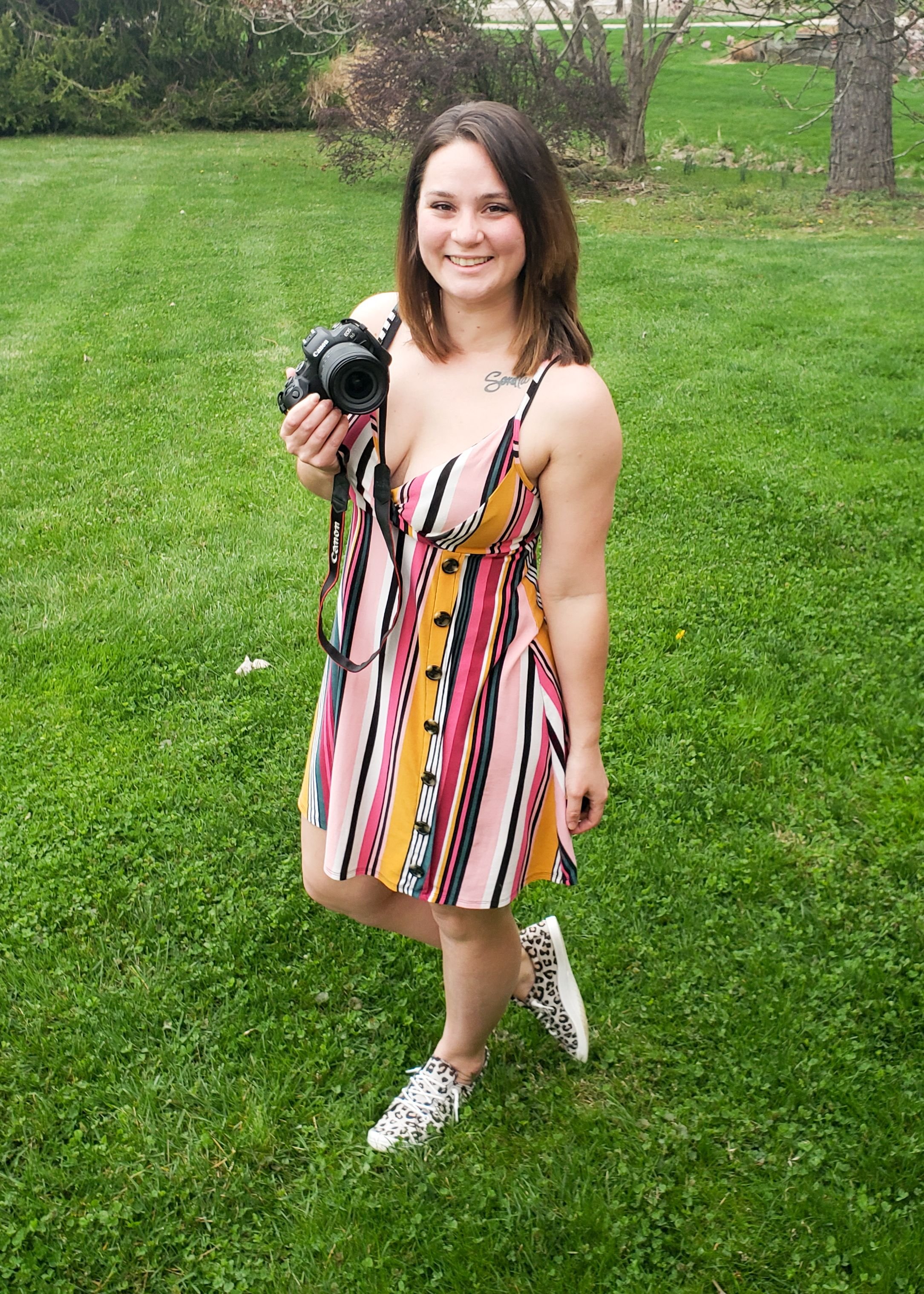 A woman standing on grass holding a camera, wearing a colorful striped dress and leopard print sneakers, with trees and greenery in the background.