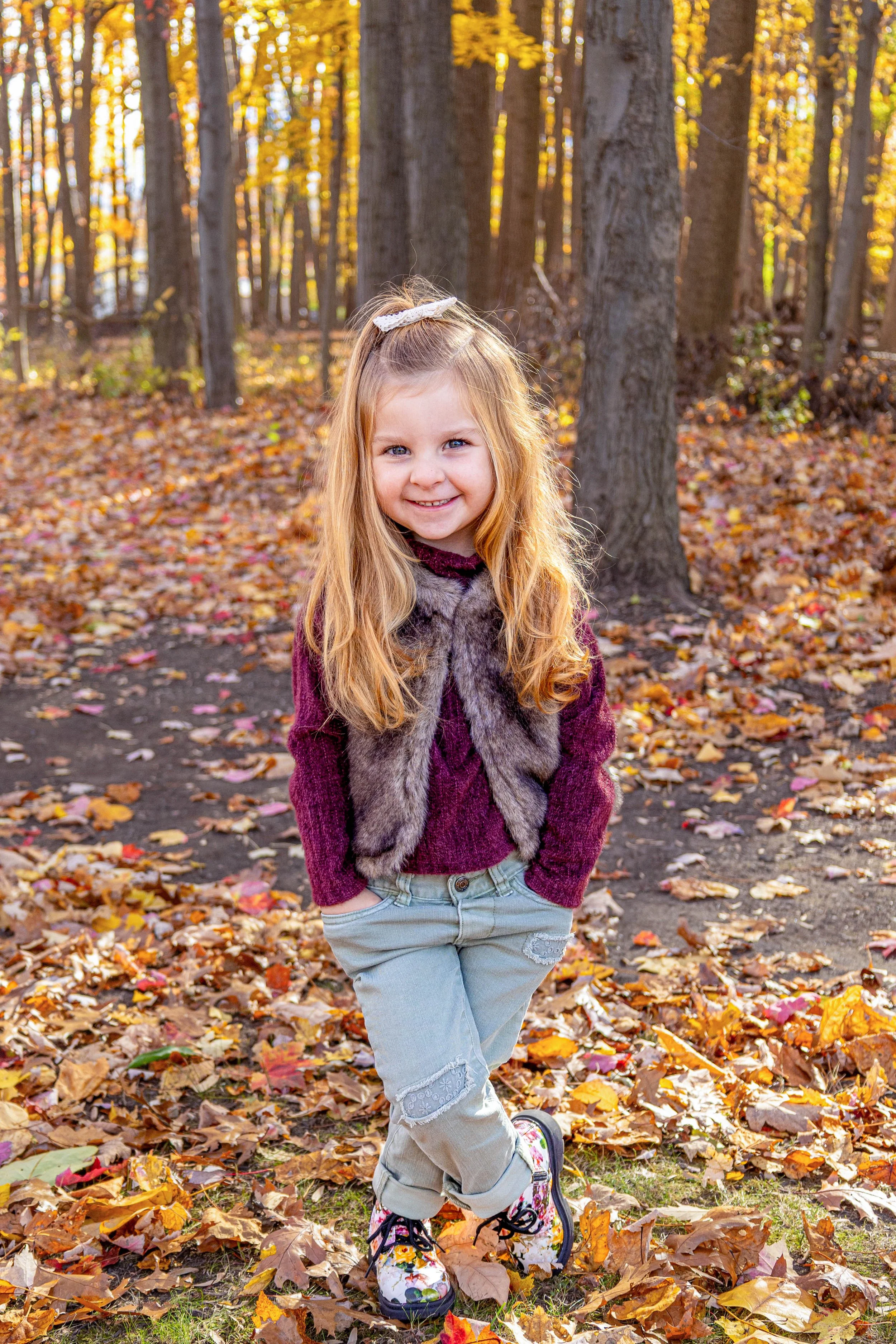 A young girl with long blonde hair, wearing a white bow, a maroon sweater, a gray faux fur vest, and light-colored jeans, stands outdoors in an autumn forest with leaves on the ground and tall trees in the background.