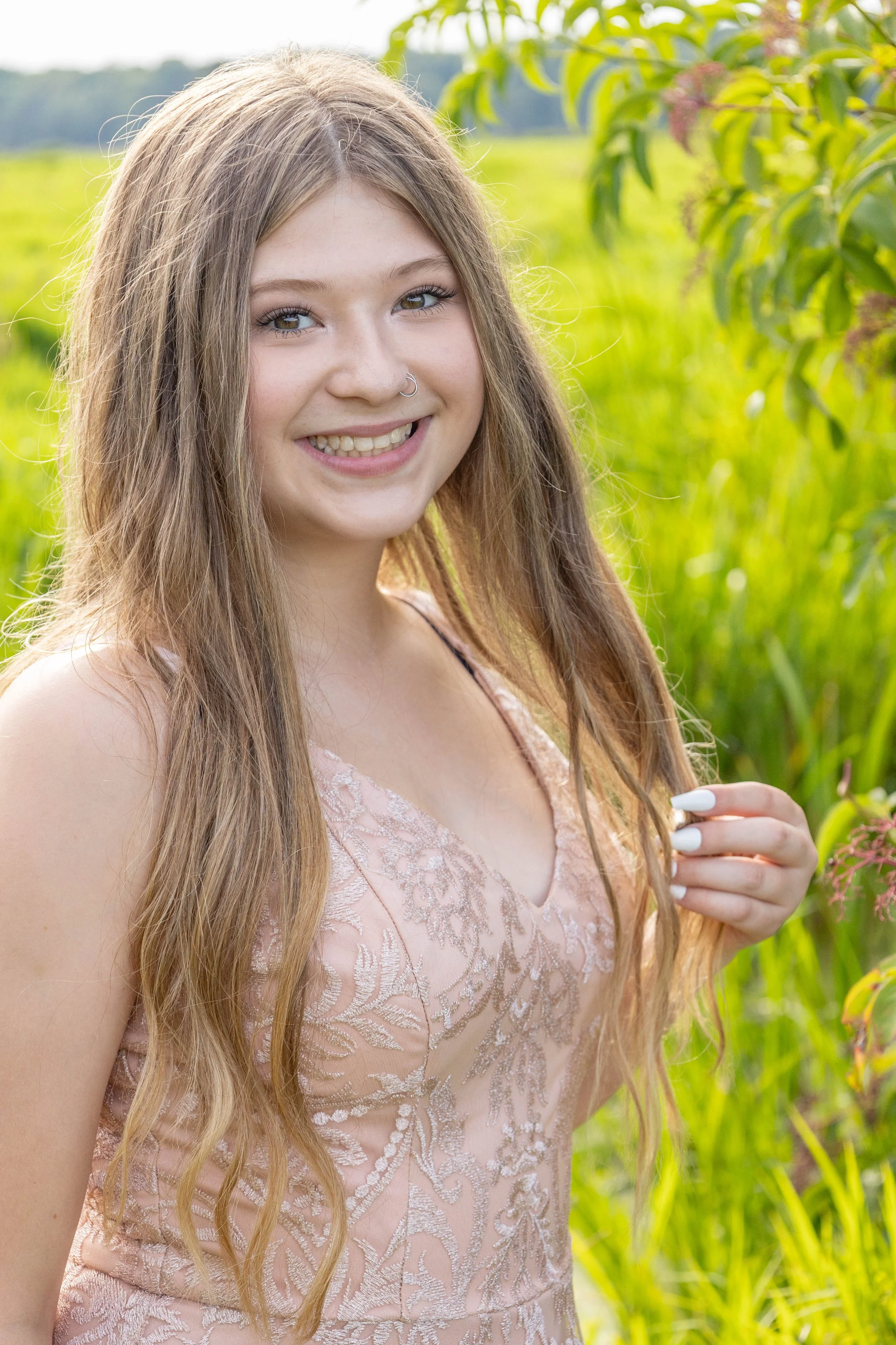 A young woman with long light brown hair, wearing a light pink dress with lace details, standing outdoors in a grassy area with green plants. She is smiling and has a nose ring.