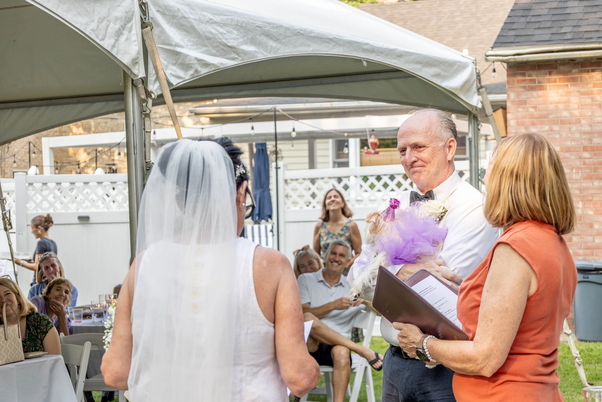 A wedding ceremony taking place in a backyard under a white canopy, with the bride and groom facing each other. Guests are seated, smiling, and watching the ceremony. The officiant, holding a folder, is speaking to the couple.