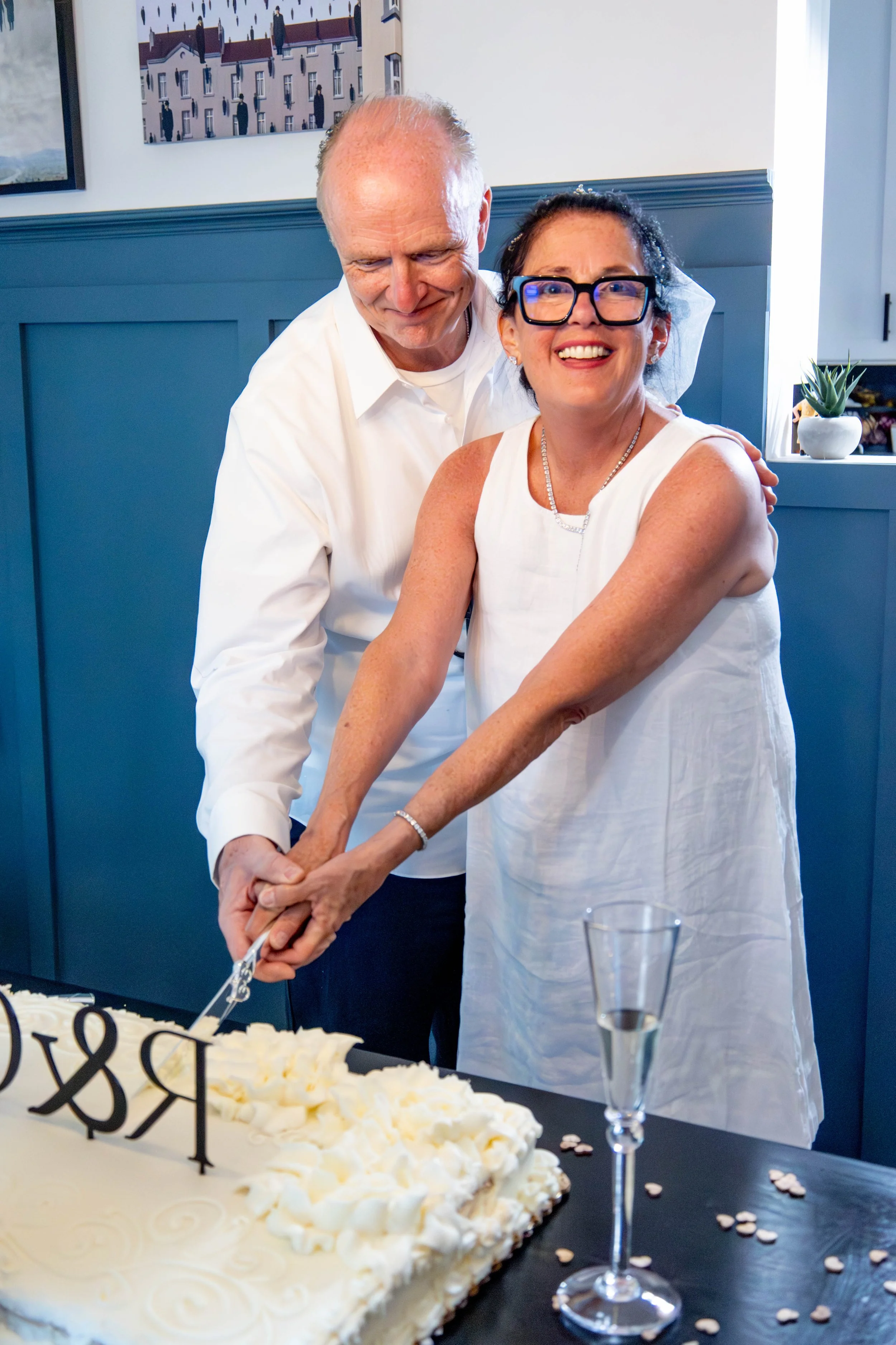 A couple cutting a wedding cake together, with a large white cake decorated with white swirls and a 'LOVE' topper, and a woman wearing glasses and a white dress. The setting appears to be a celebration or wedding reception with a blue wall, framed pi