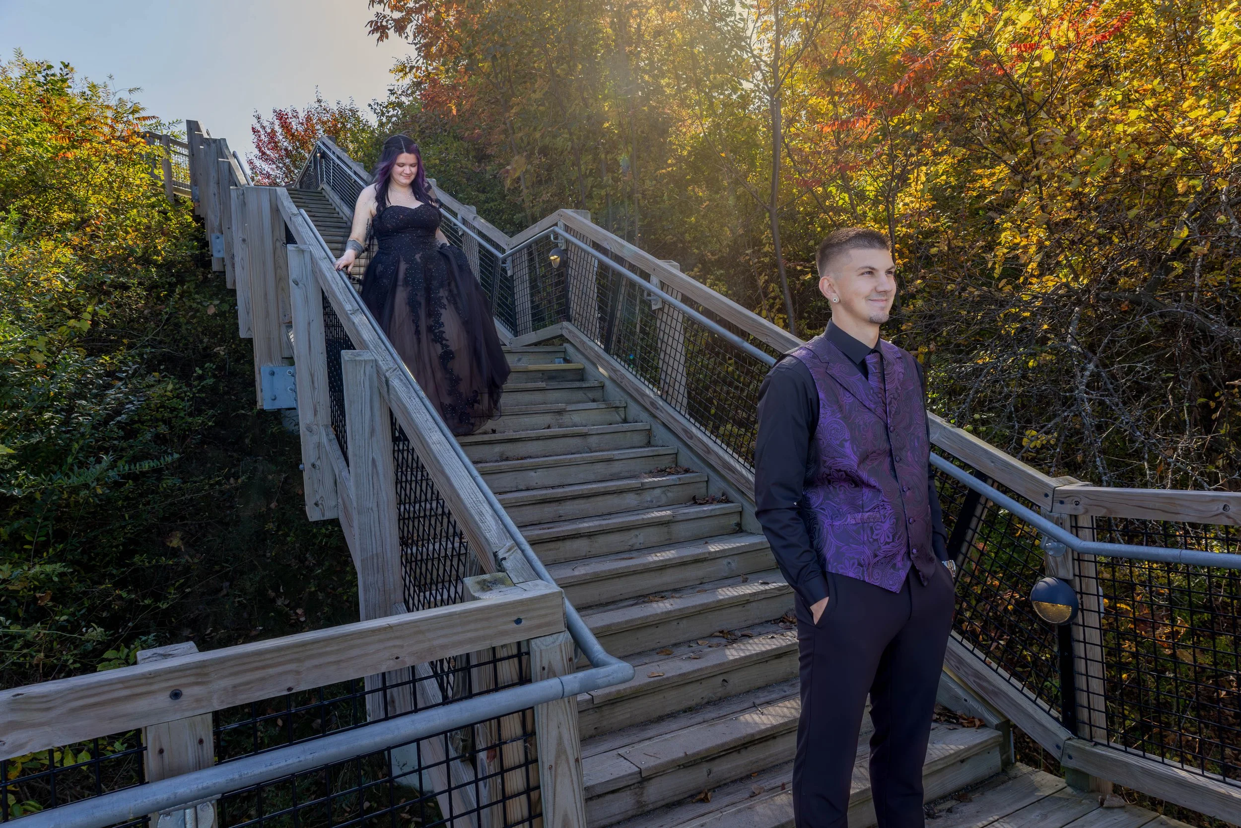 A young man in a purple vest and black shirt standing at the bottom of a wooden outdoor staircase, with a young woman in a black gown descending the stairs amid trees with autumn leaves.