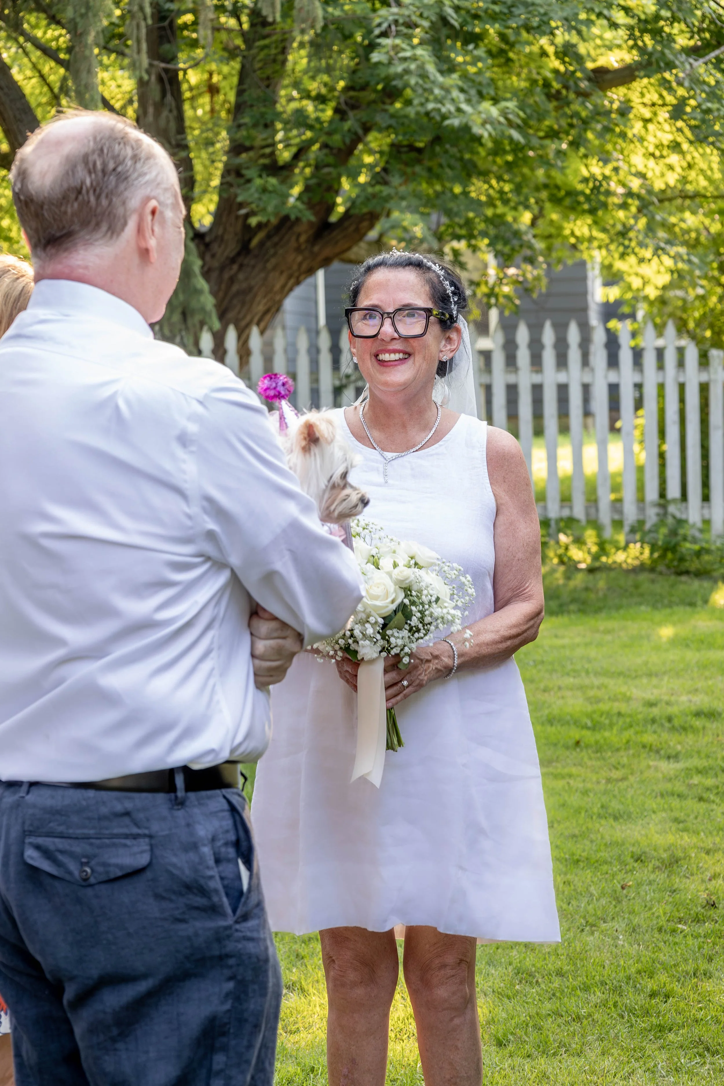 A woman in a white dress holding a bouquet of white roses, smiling and looking at a man holding a small dog with pink flowers attached. The woman wears glasses and a necklace, and they are outdoors near a white picket fence and green trees.
