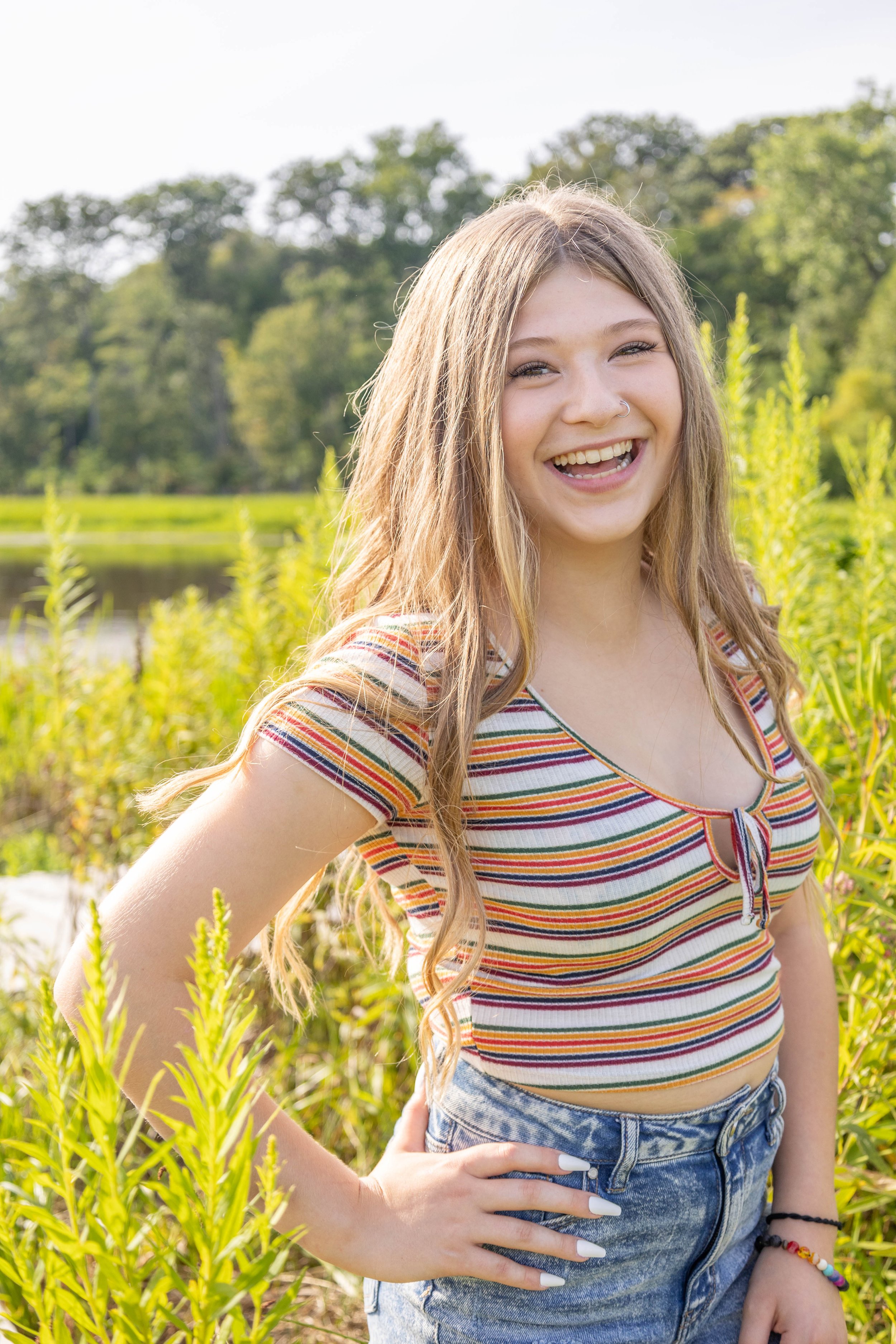 A young woman with long, wavy brown hair smiling outdoors in a natural setting with trees and tall grass, wearing a colorful striped t-shirt and high-waisted denim shorts.