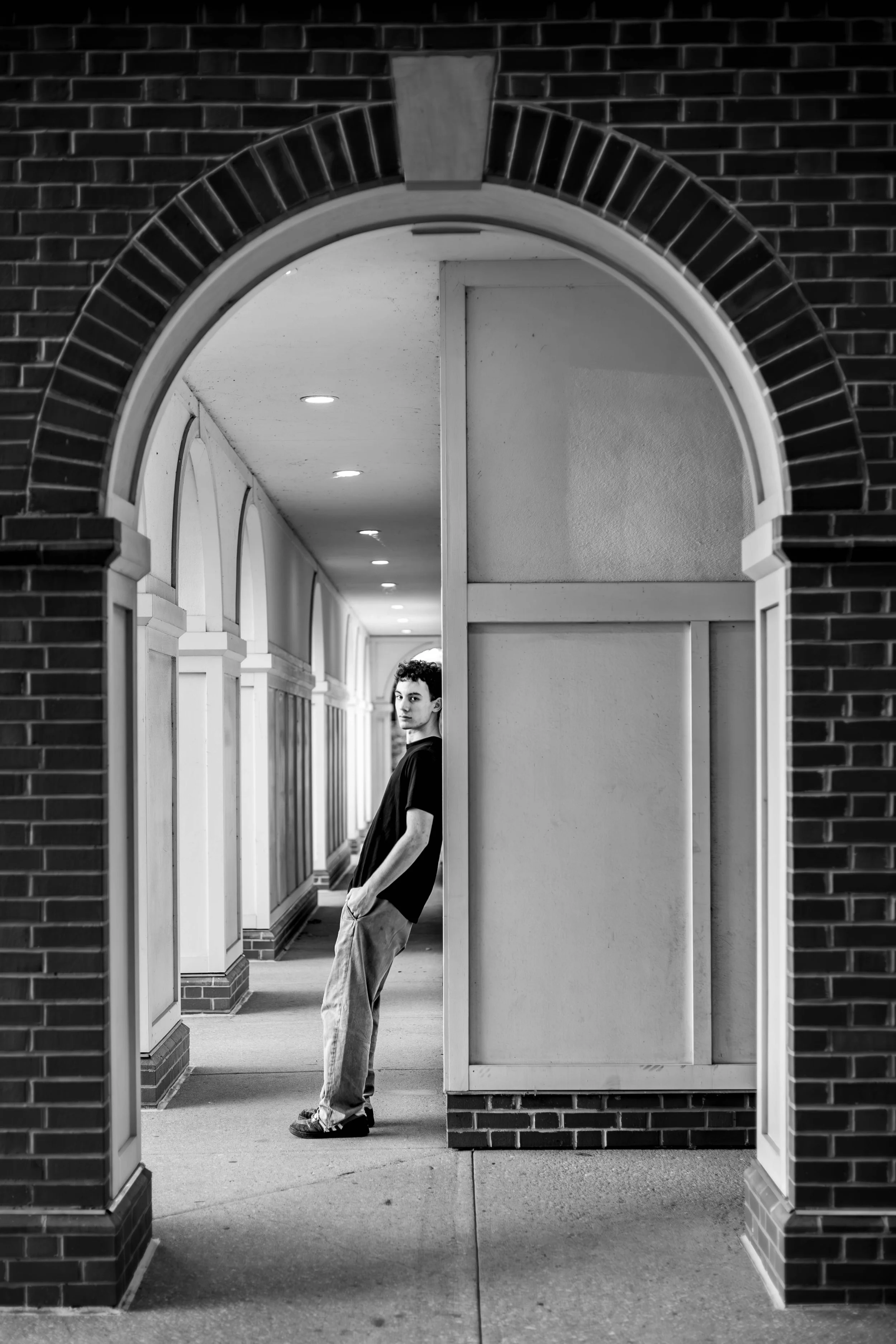 A young man in black shirt and light colored pants leaning against a low wall in an arched hallway with brick framing