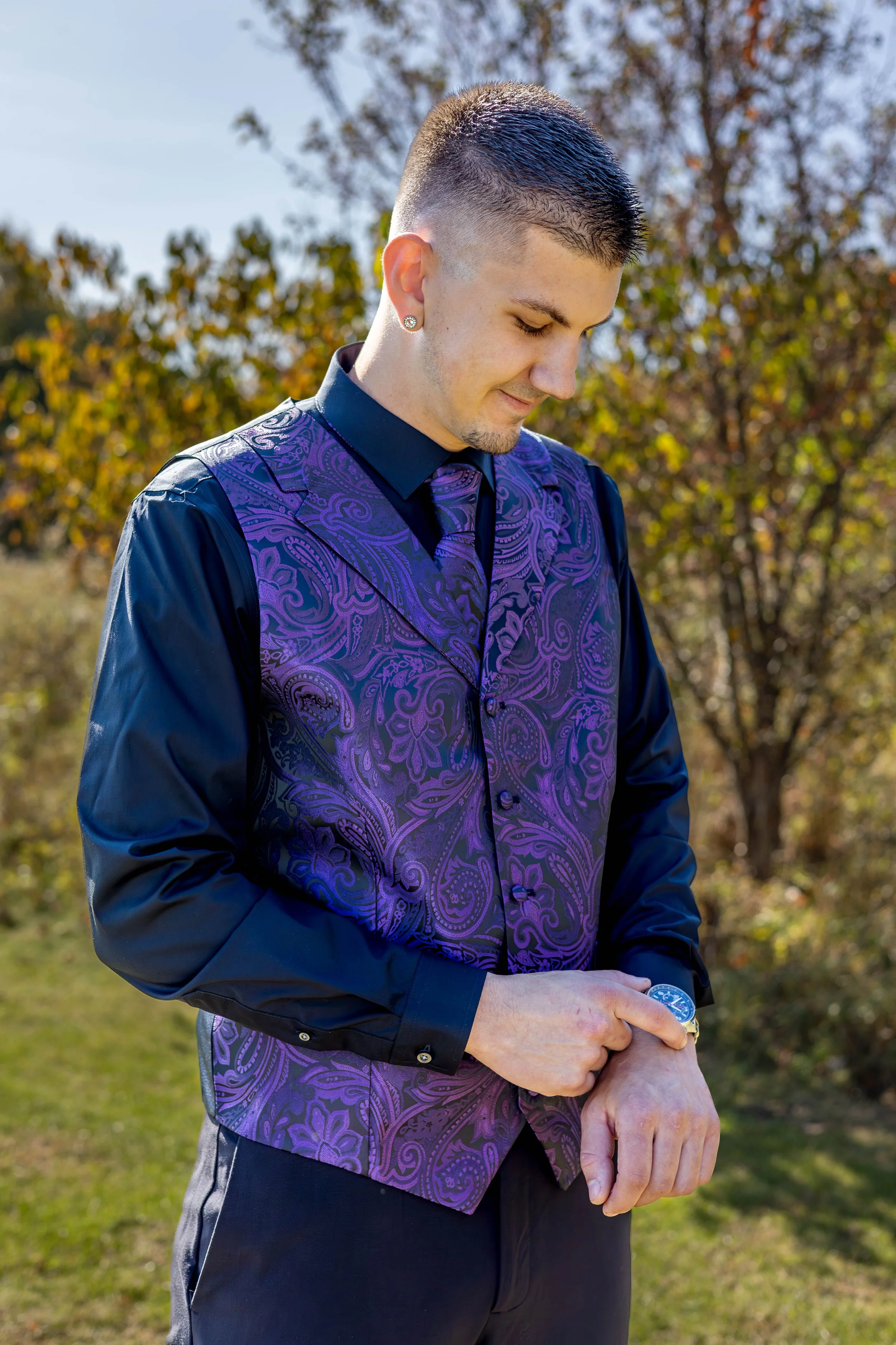 Young man with short, dark hair wearing a purple paisley vest over a black shirt, standing outdoors with trees in the background, looking down with a slight smile, holding a handshake with someone.