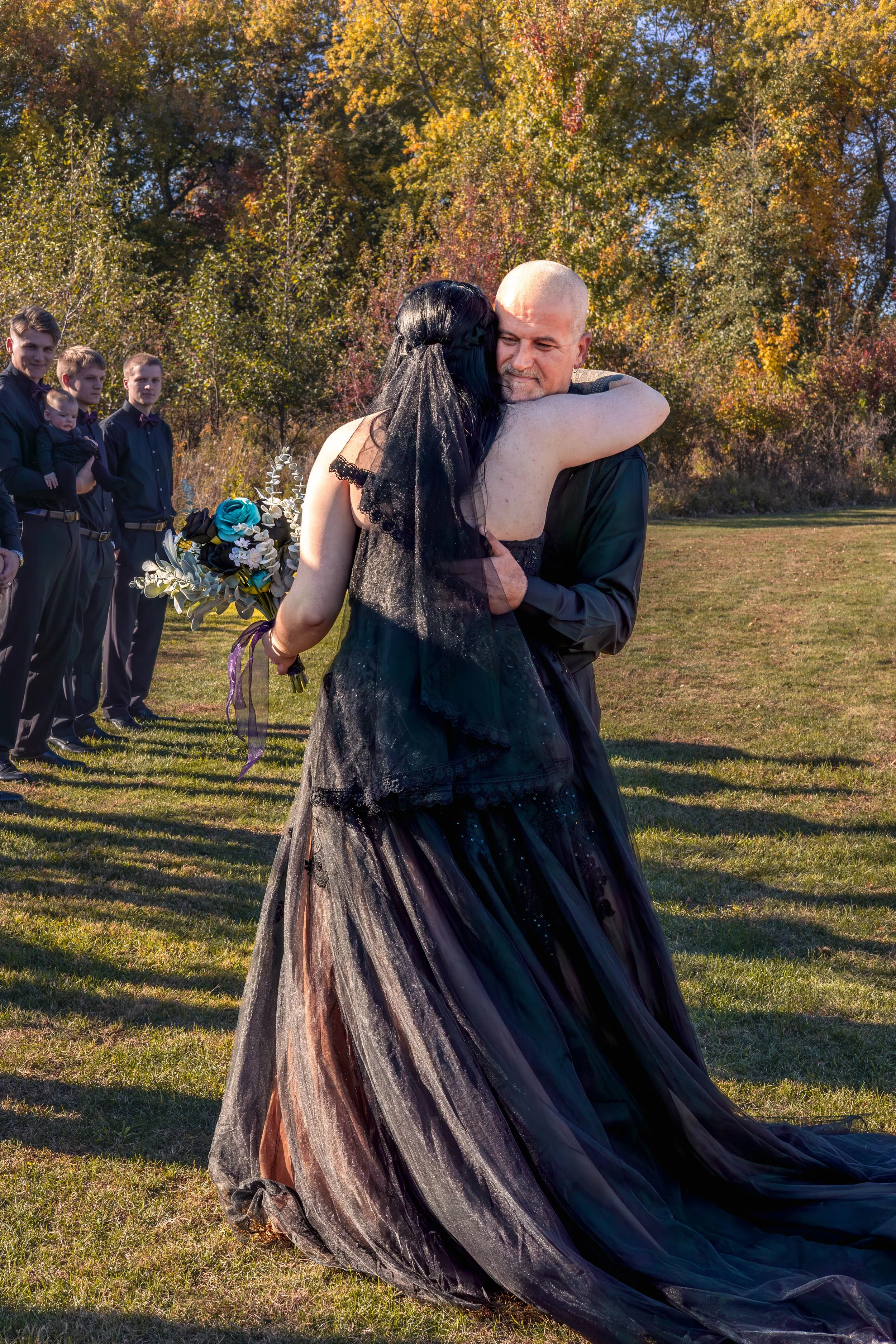 A couple embracing during a wedding ceremony outdoors with friends standing in the background, autumn trees in the distance.