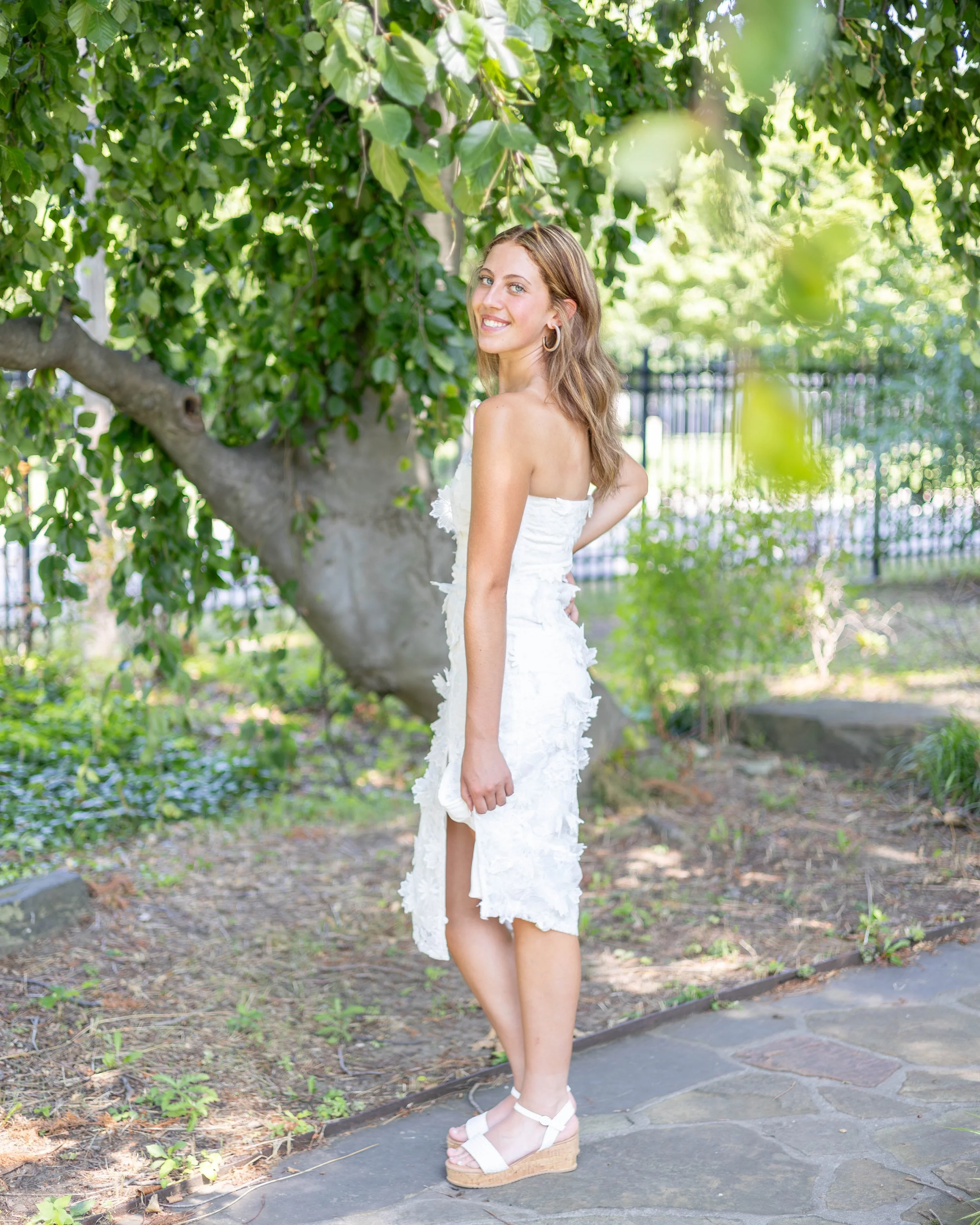 A young woman with long, wavy brown hair and blue eyes stands outdoors in front of a leafy green tree, smiling and looking over her shoulder at the camera. She is wearing a white strapless dress with ruffled details and white wedge sandals with ankle straps. The ground is paved with stones and there is a fence in the background.