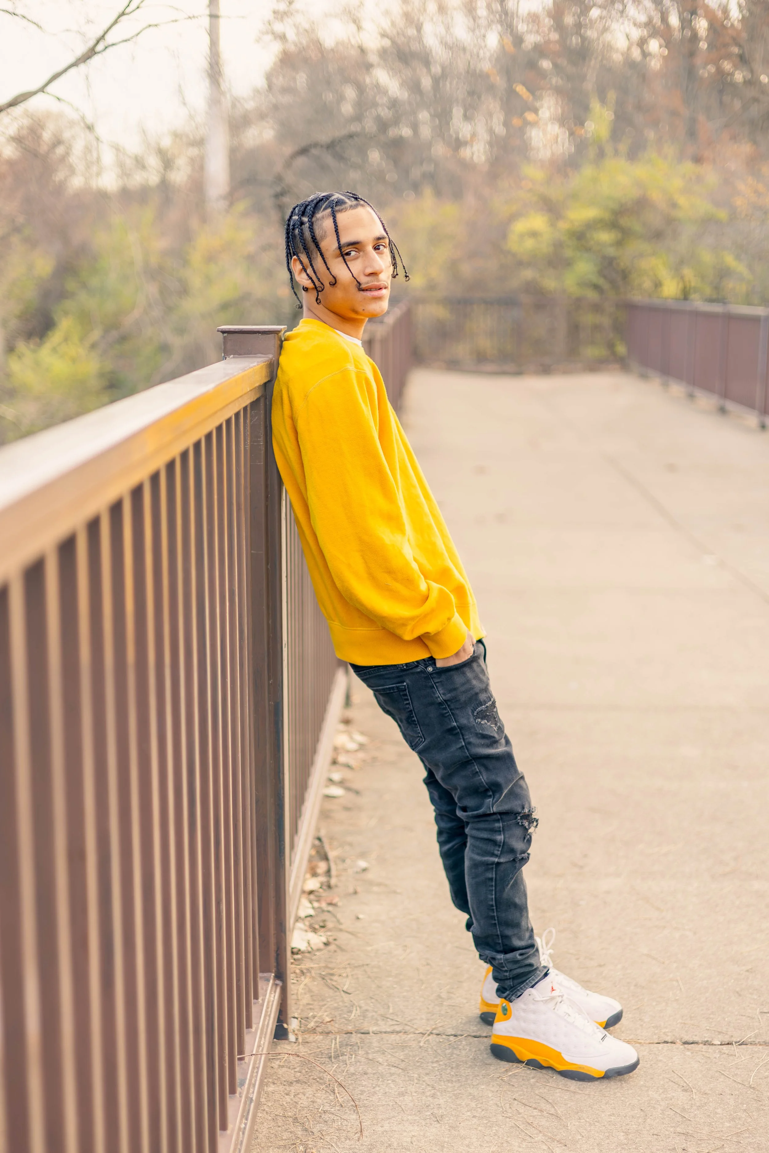 A young man leaning against a brown metal railing on an outdoor sidewalk, wearing a yellow sweatshirt, ripped black jeans, and white sneakers with yellow and black accents, in a park during autumn.