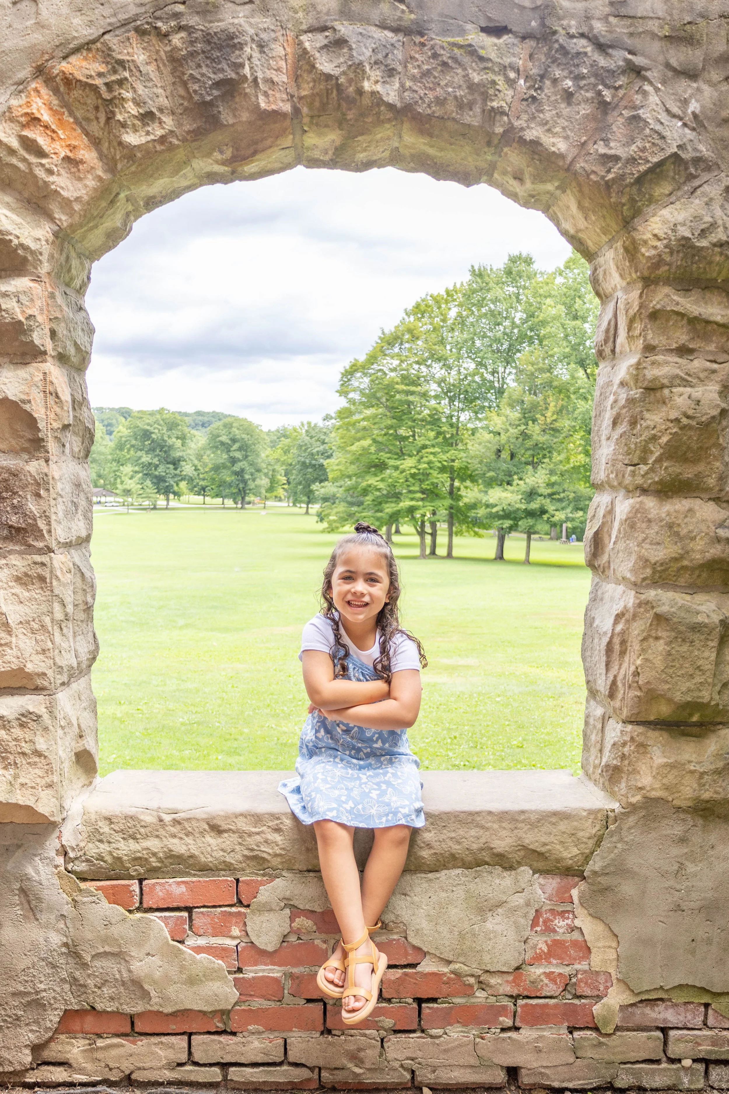 Young girl sitting on a windowsill with a stone arch, smiling, outdoors with green grass and trees in the background.