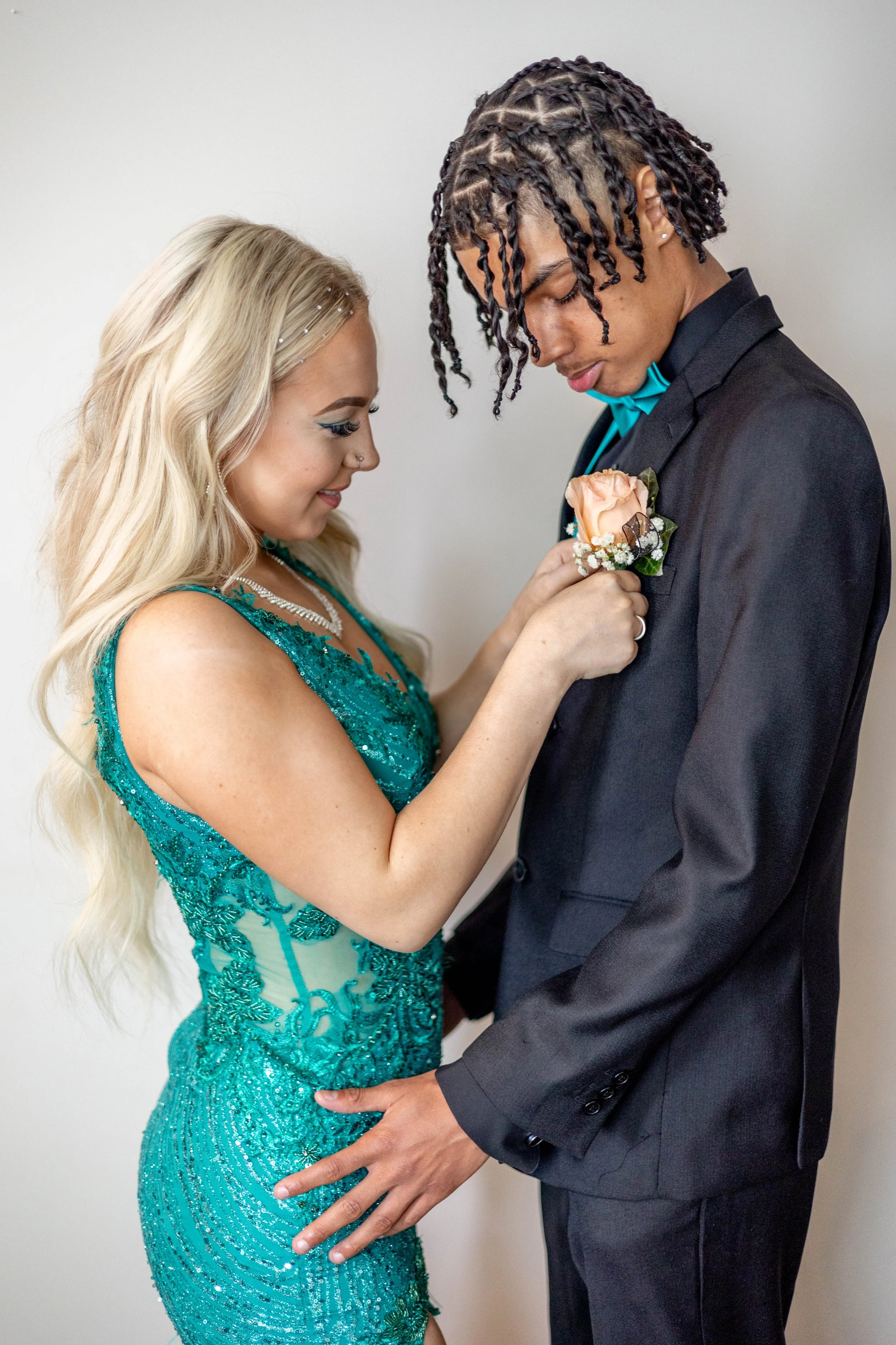 A woman in a teal, sequined gown with long blonde hair is placing a corsage on a man's suit jacket. The man has short, styled hair and is wearing a dark suit with a bow tie.